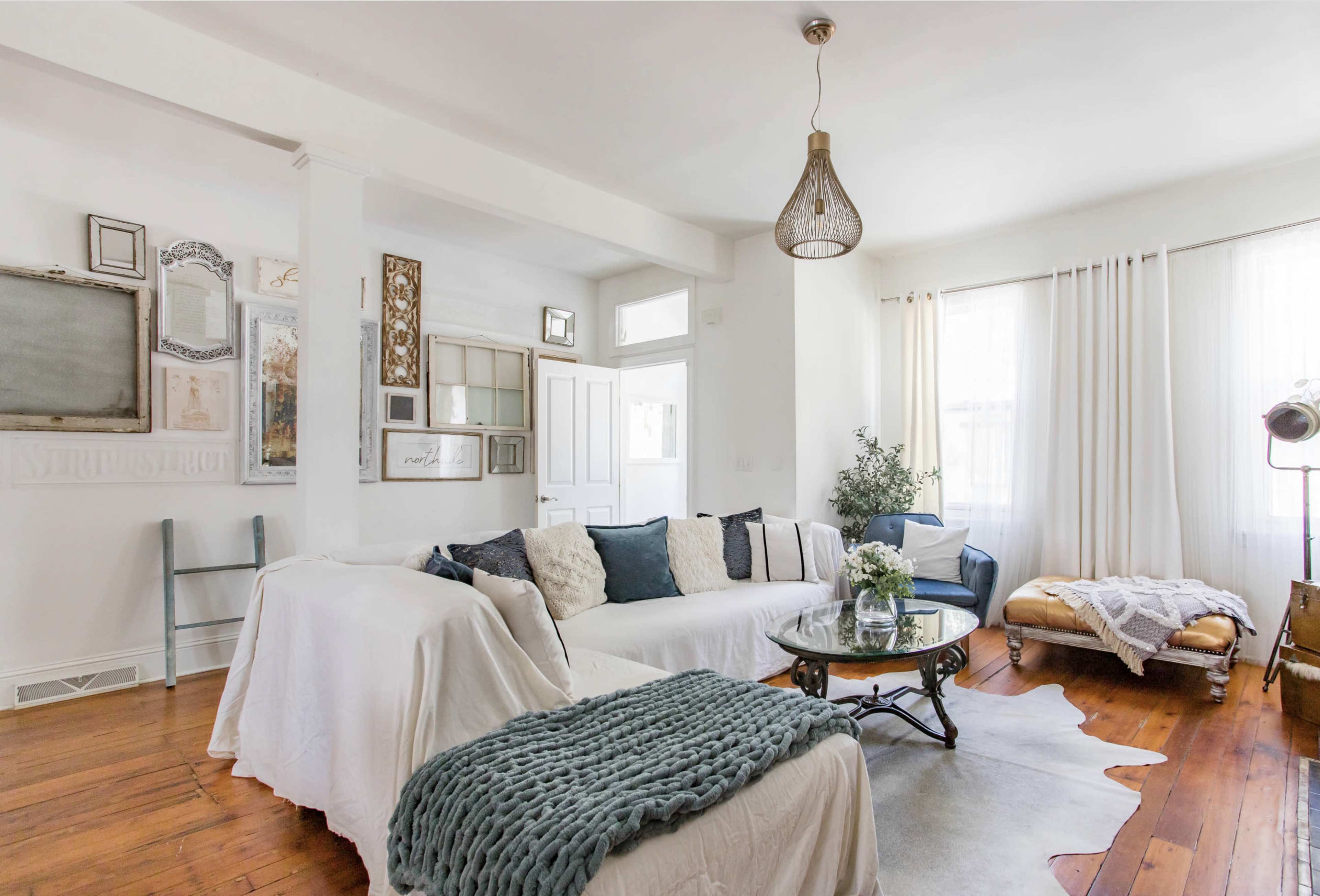 The living room features a mix of furniture styles, including a white slipcovered sofa, a glass-top coffee table, and a vintage armchair, all set against light-colored walls and hardwood flooring.