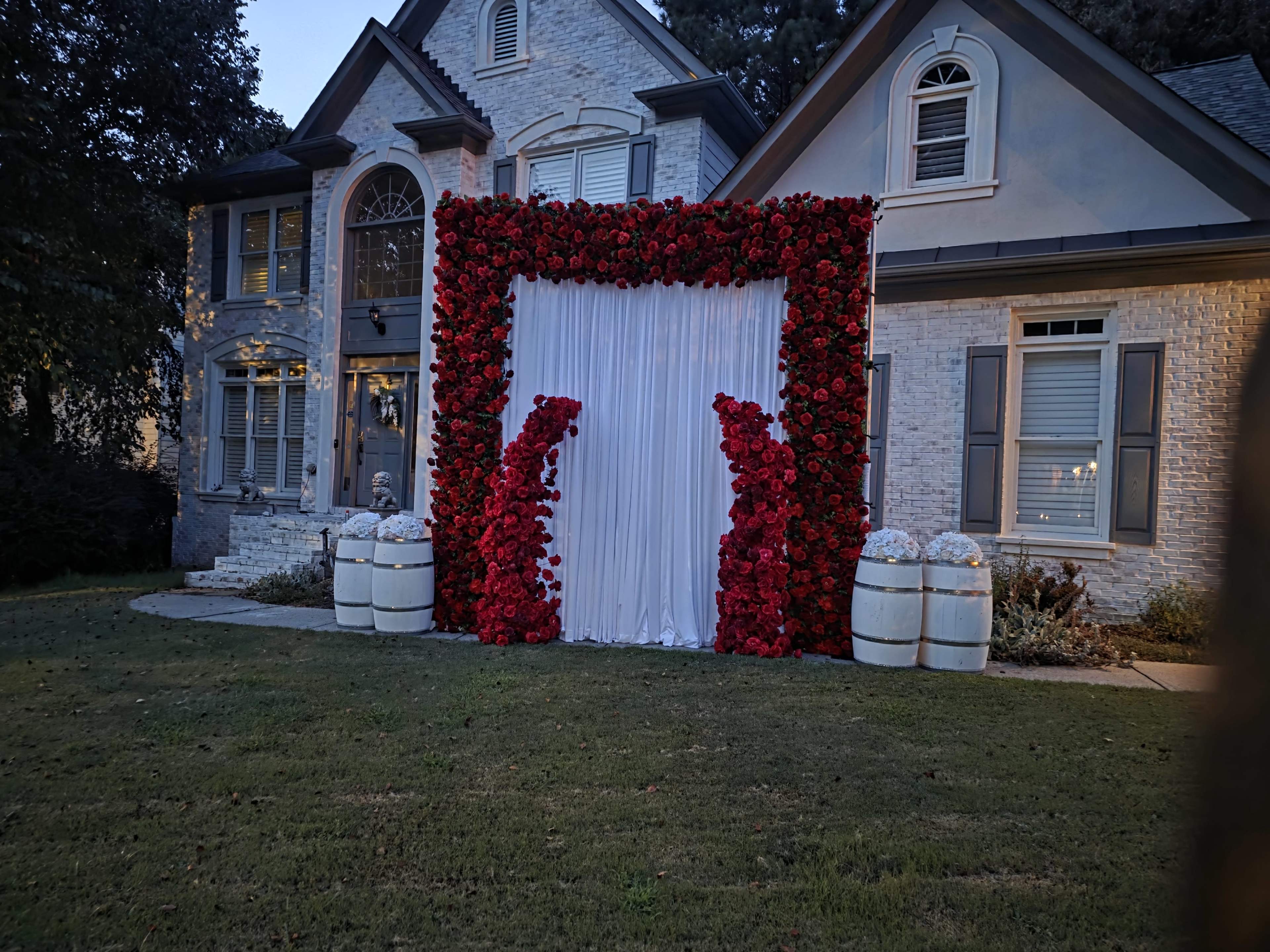A decorative floral arch made of red roses and white drapery is set up in front of a large, two-story house.