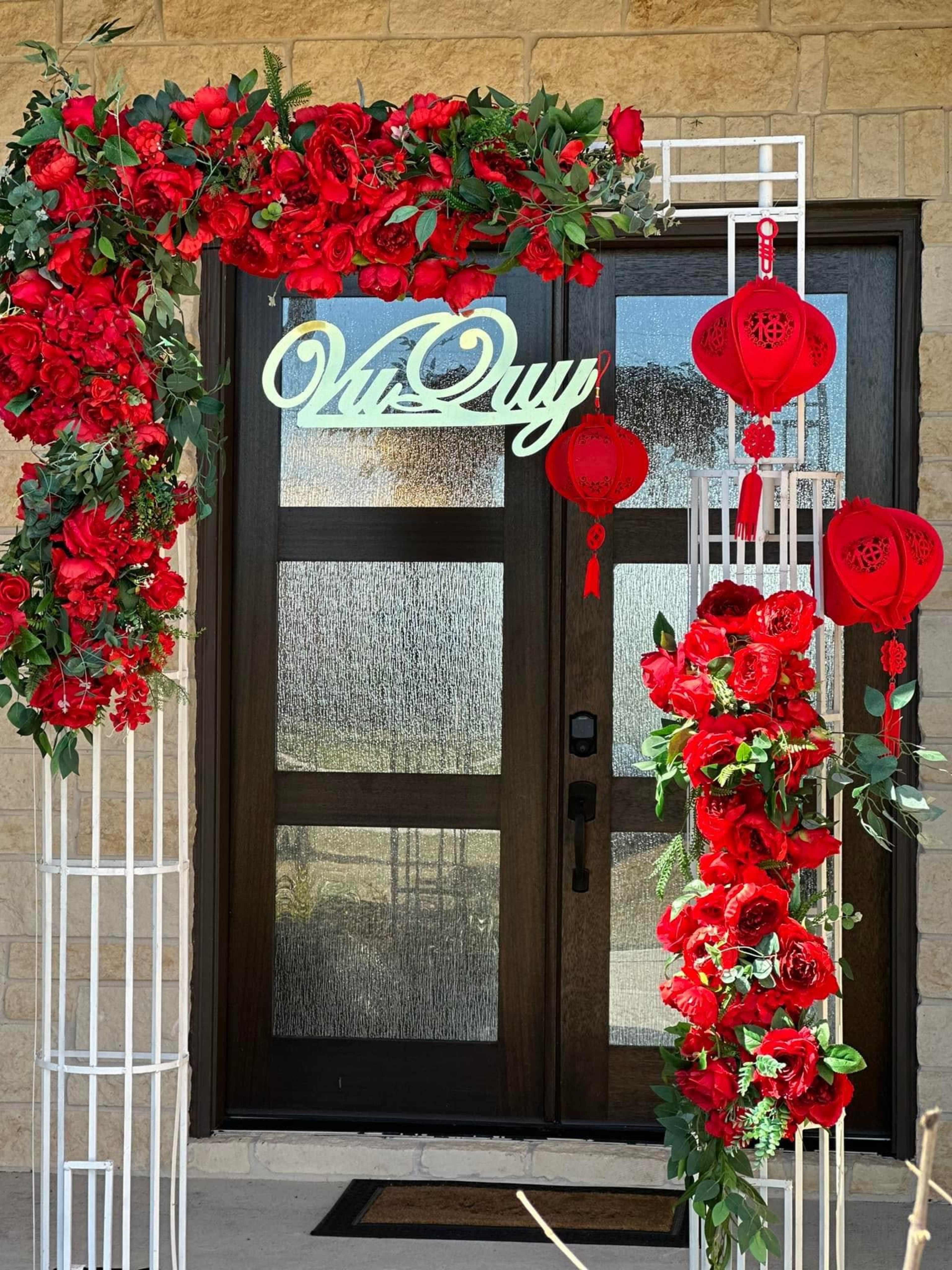A decorative arch adorned with red flowers and lanterns stands at the entrance of a house with a wooden door.