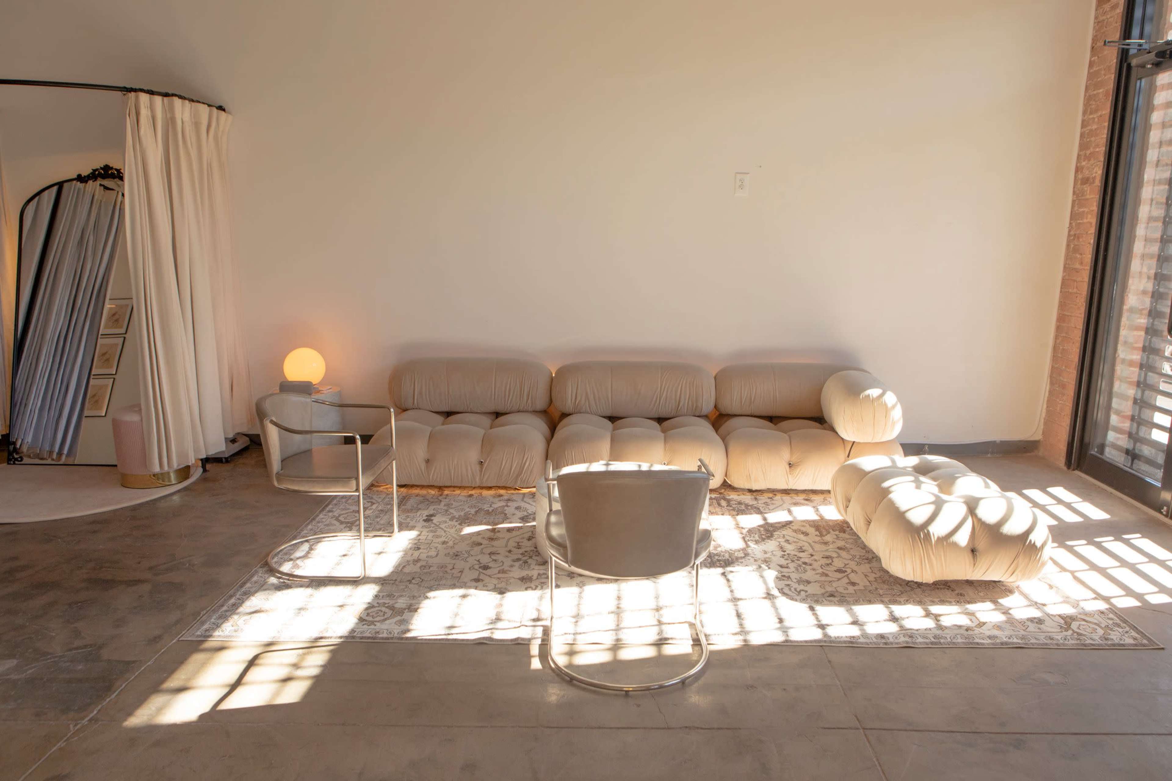 The image shows a spacious room with a light-colored, plush sectional sofa, a minimalist chair, and a round floor lamp, all arranged on a decorative area rug under natural light from large windows.