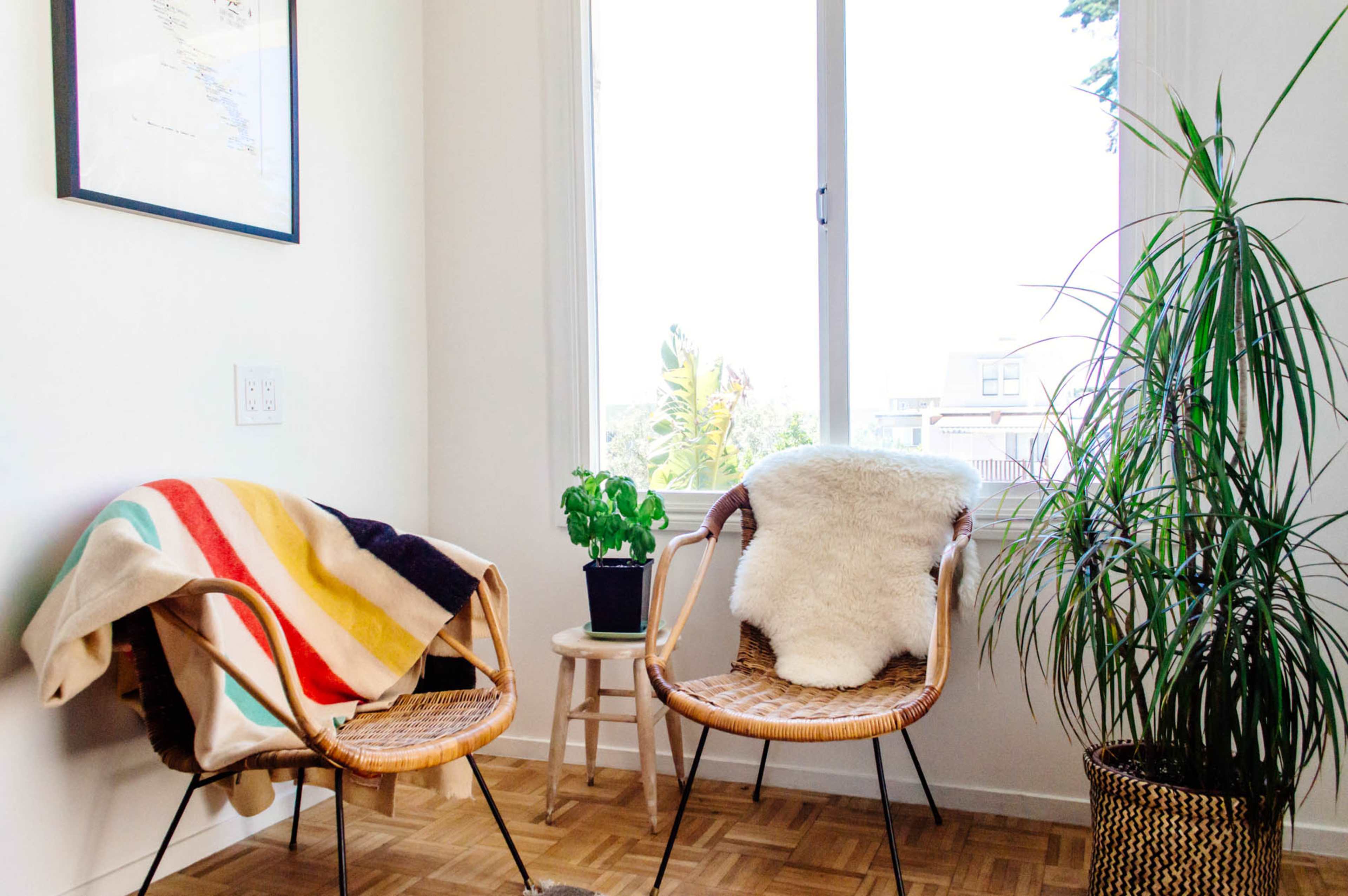 Two wicker chairs with colorful blankets and a plant sit beside a window in a bright, airy corner.