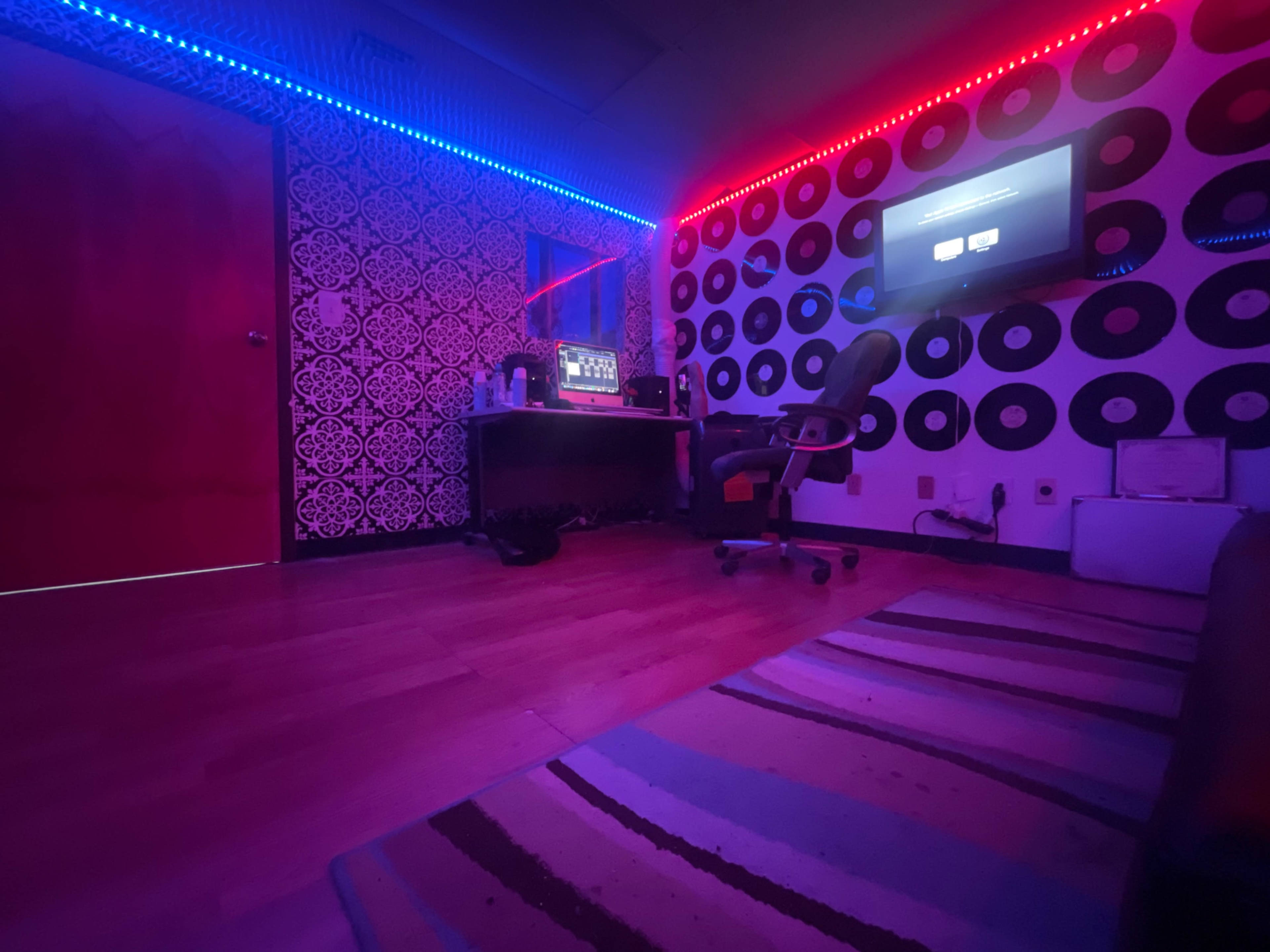 A dimly lit room with colorful LED strip lighting, a desk with a computer setup, and walls decorated with vinyl records.