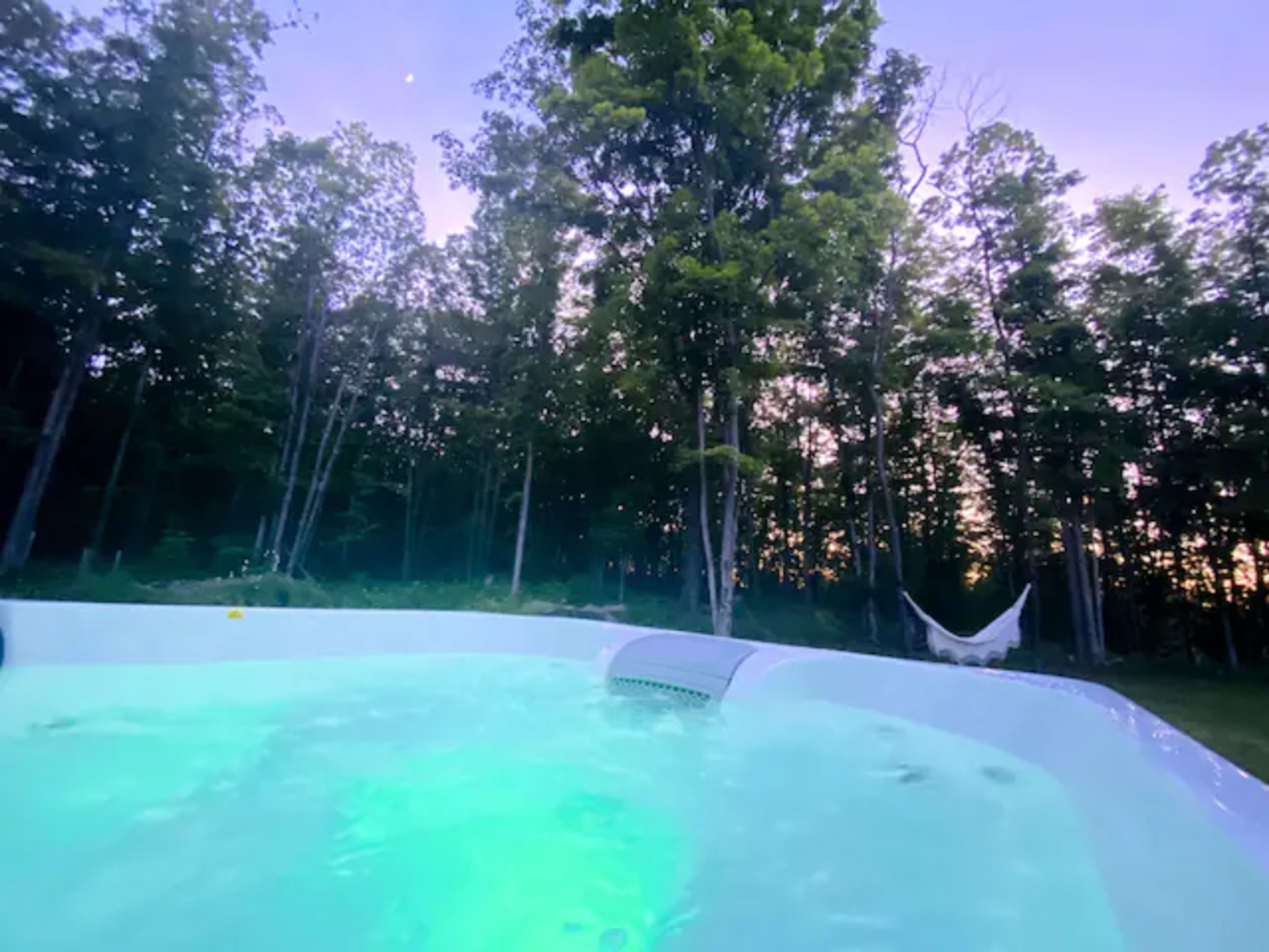 The image shows a swimming pool illuminated with green light, surrounded by trees and a hammock in a forested area during twilight.