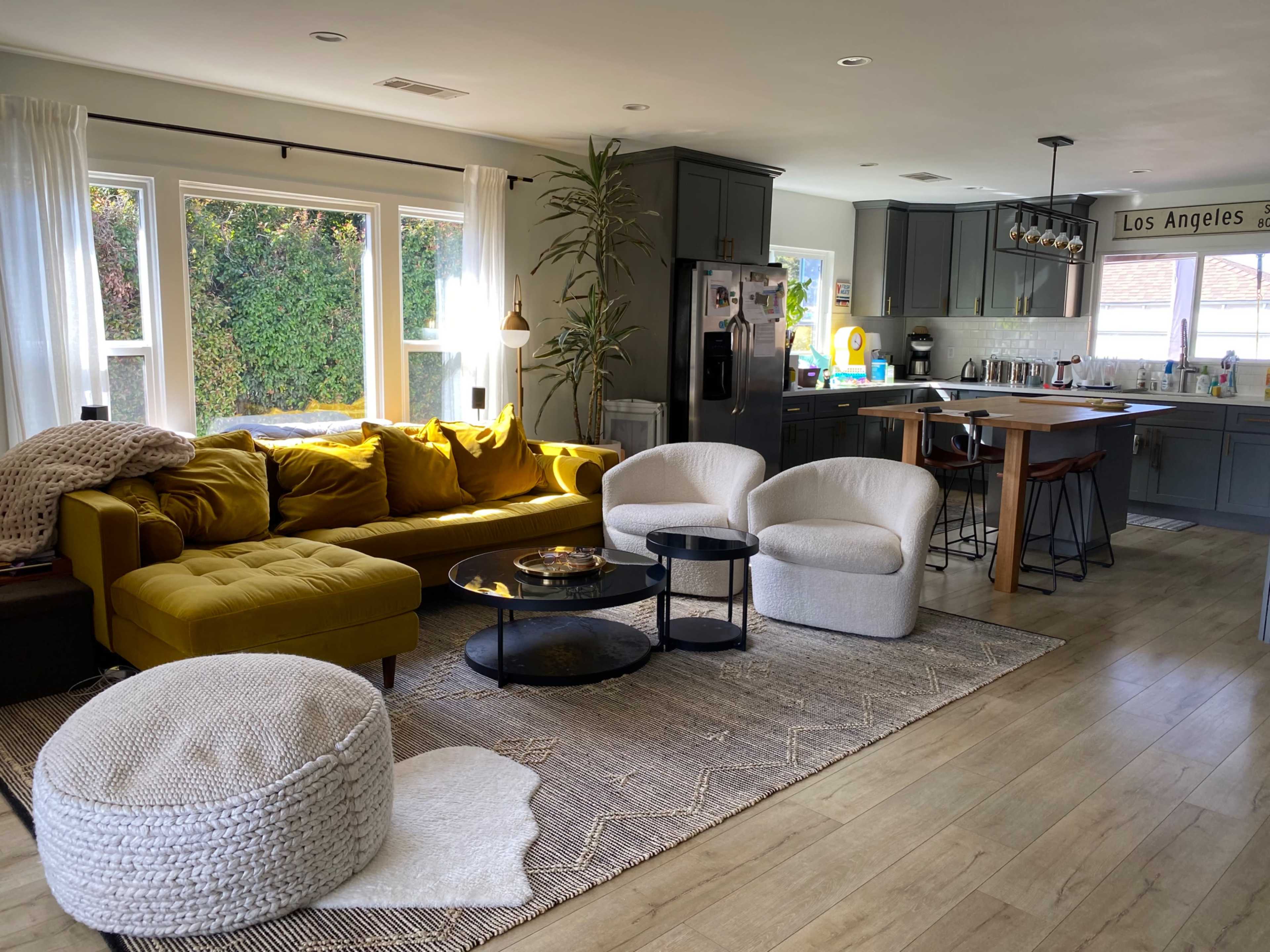 A modern living room with a yellow sectional sofa, two white chairs, a round coffee table, and a kitchen area in the background.