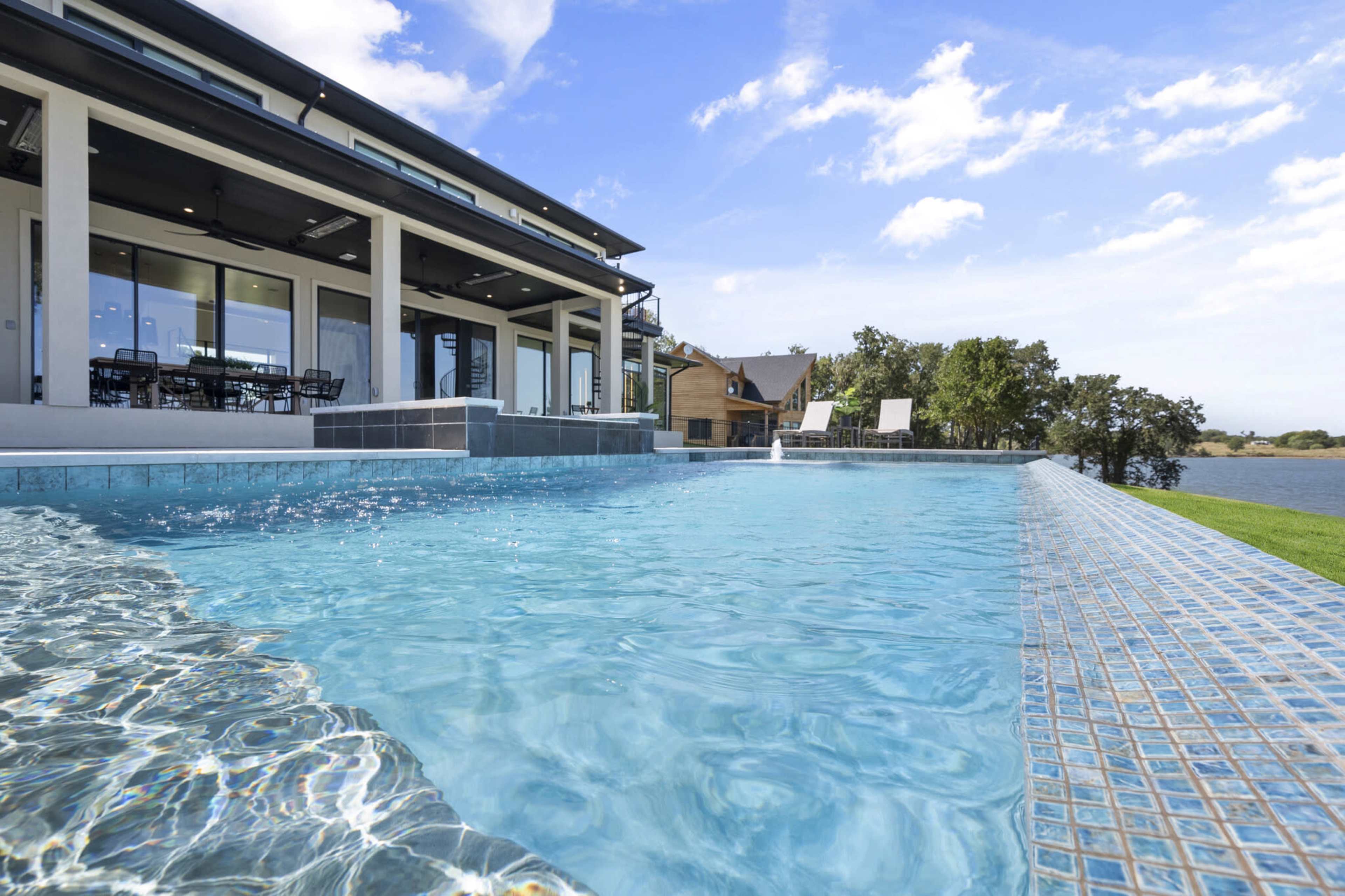 A modern house with large windows overlooks a swimming pool that extends to the edge of a body of water.