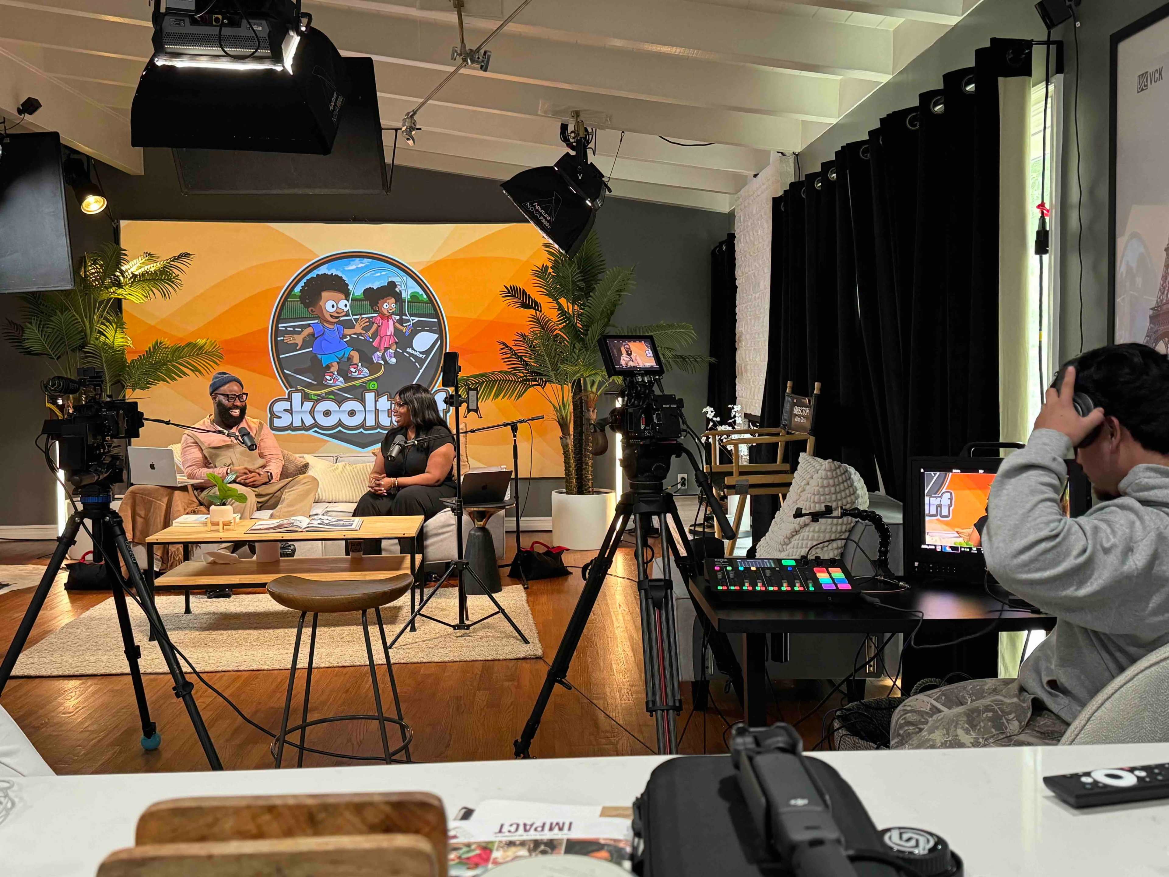The image shows a studio setup with two hosts sitting on a couch and cameras positioned around them, featuring a colorful backdrop with the "skoolit" logo.