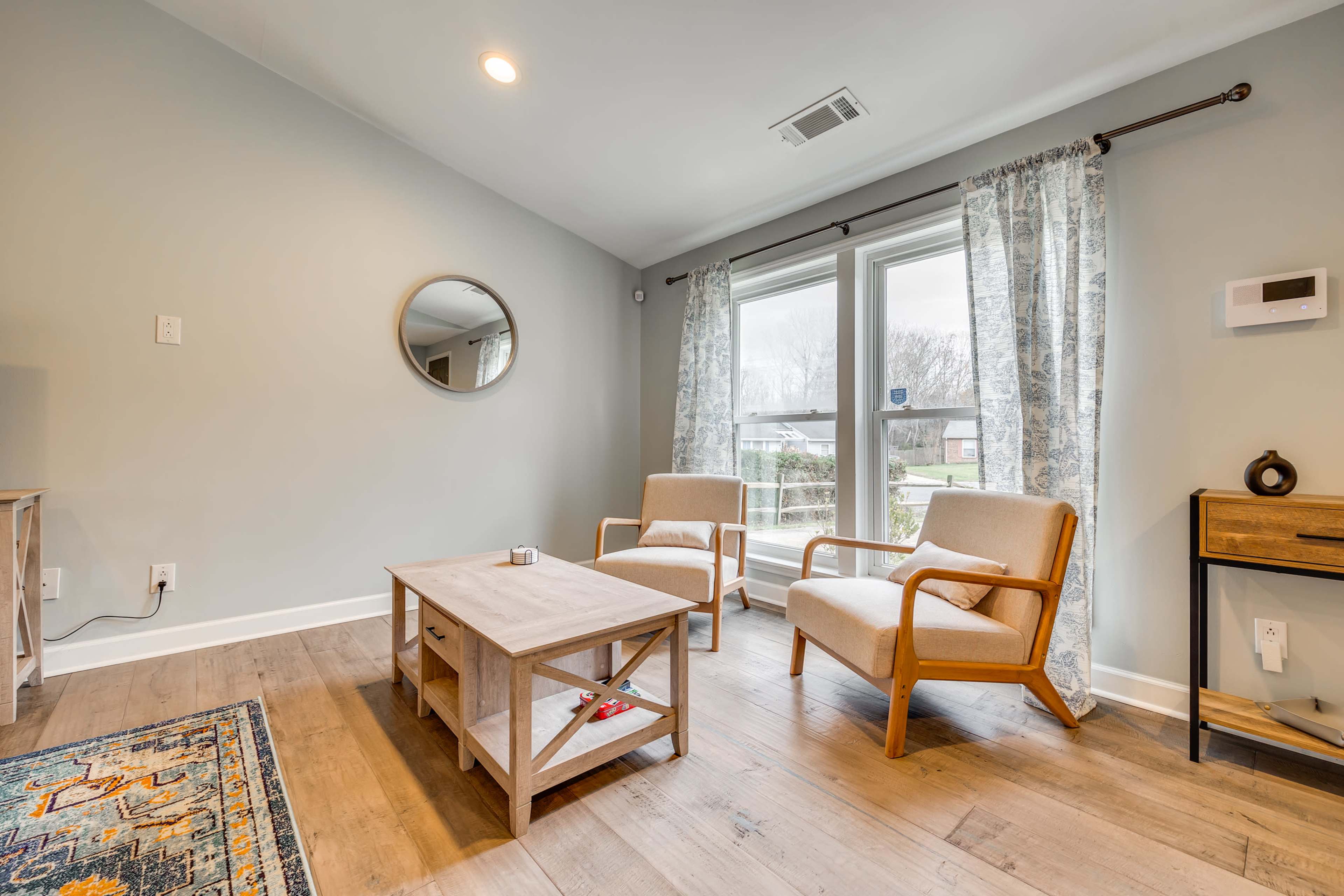 A living room with two wooden chairs, a neutral coffee table, a round mirror on the wall, and large windows with curtains, allowing natural light to enter.