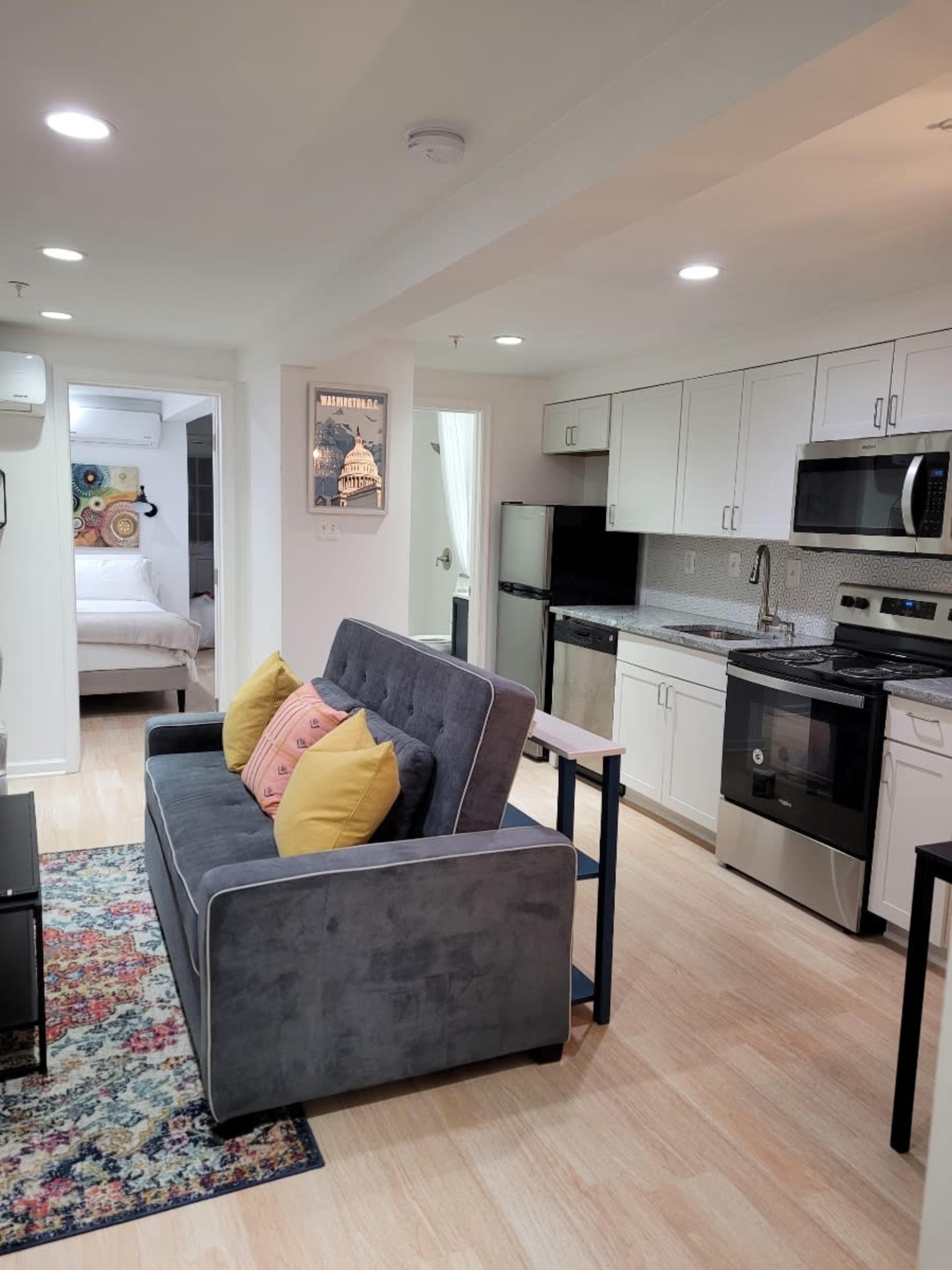 A modern kitchenette and living area feature a gray sofa with yellow pillows and a dining table in a well-lit, open space.