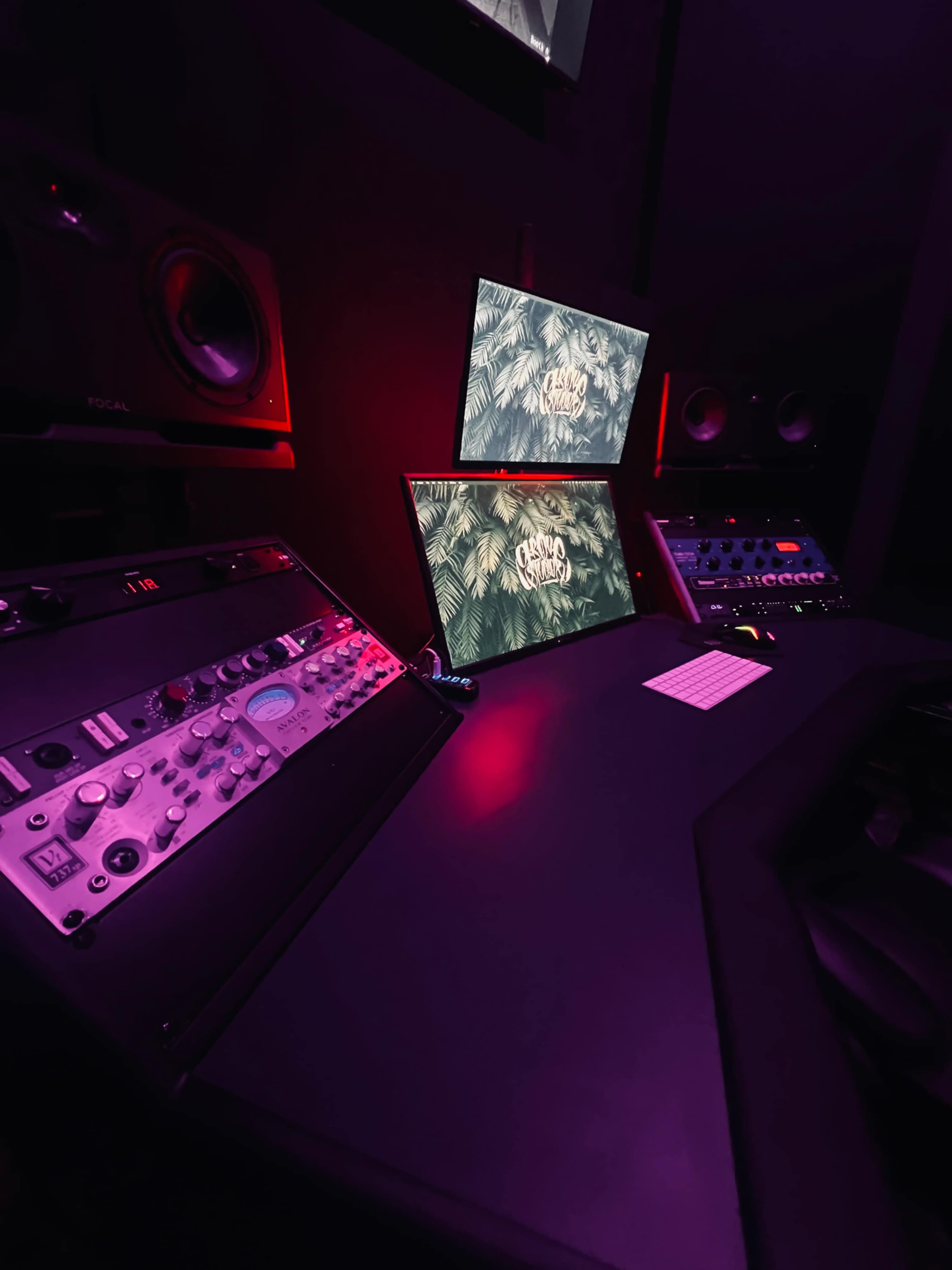 The image shows a modern music production studio with two monitors, a mixing console, and audio equipment, illuminated by soft purple lighting.