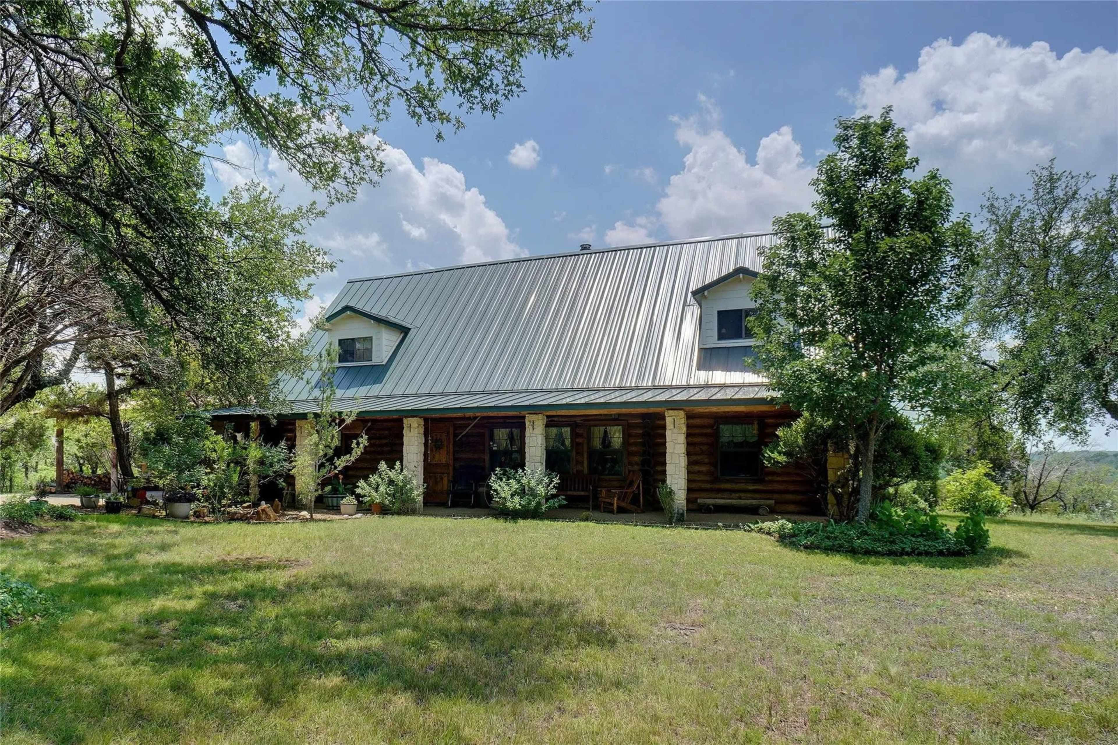 A rustic log cabin with a metal roof is surrounded by greenery and trees under a partly cloudy sky.
