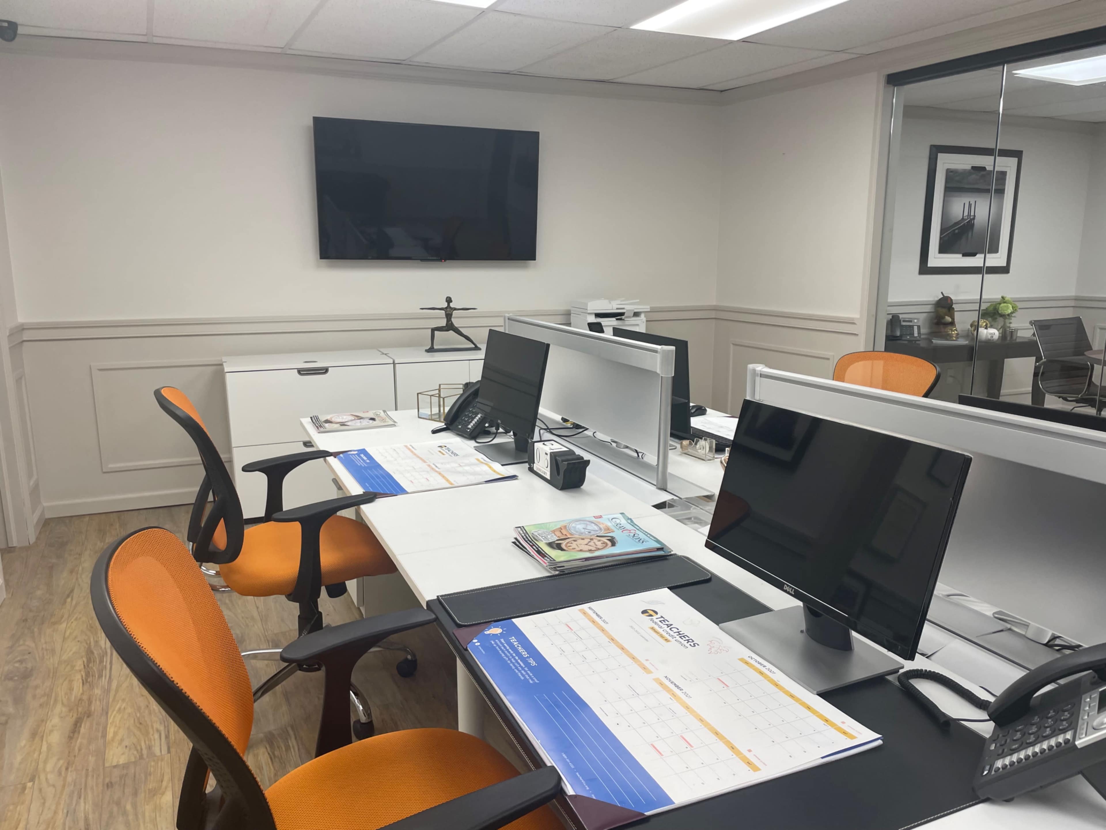 A modern office space with two orange swivel chairs and two computer workstations, along with a television mounted on the wall.