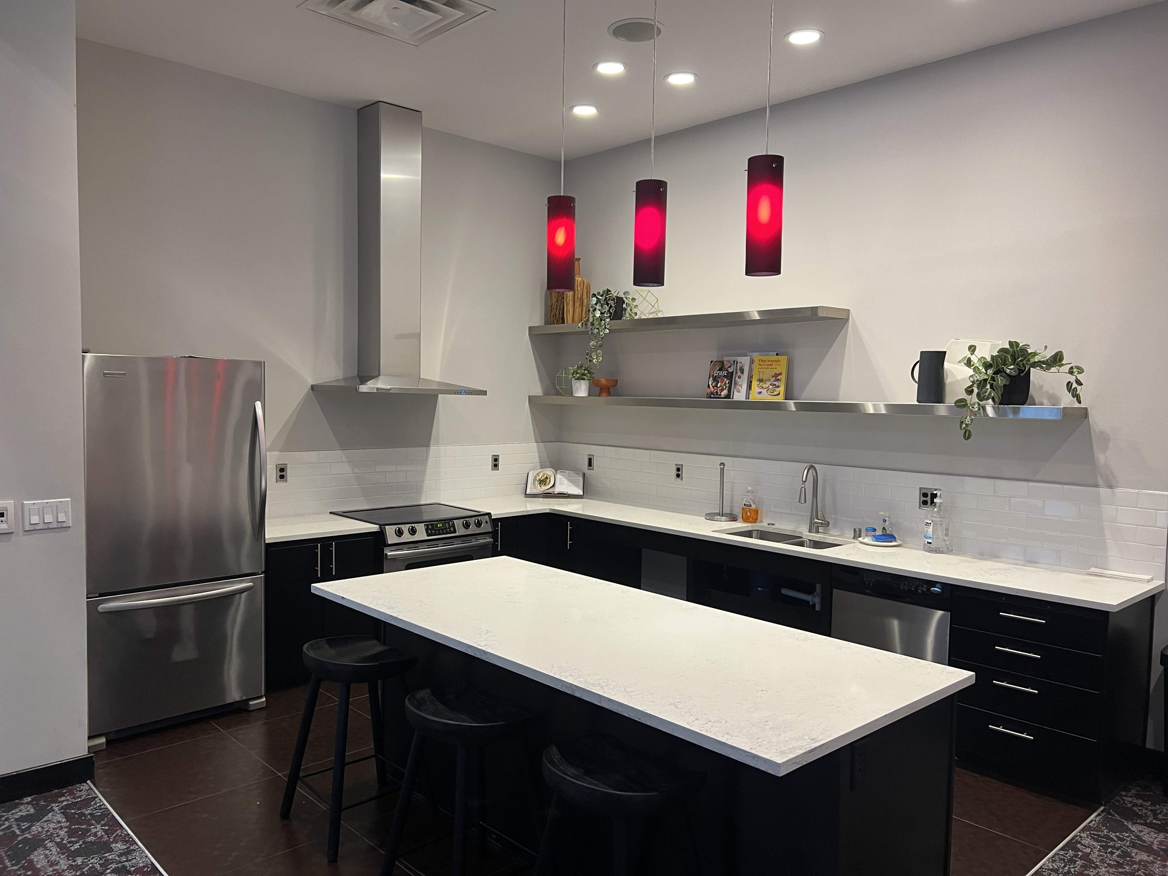 A modern kitchen featuring stainless steel appliances, dark cabinetry, and a large island with bar stools, illuminated by red pendant lights.
