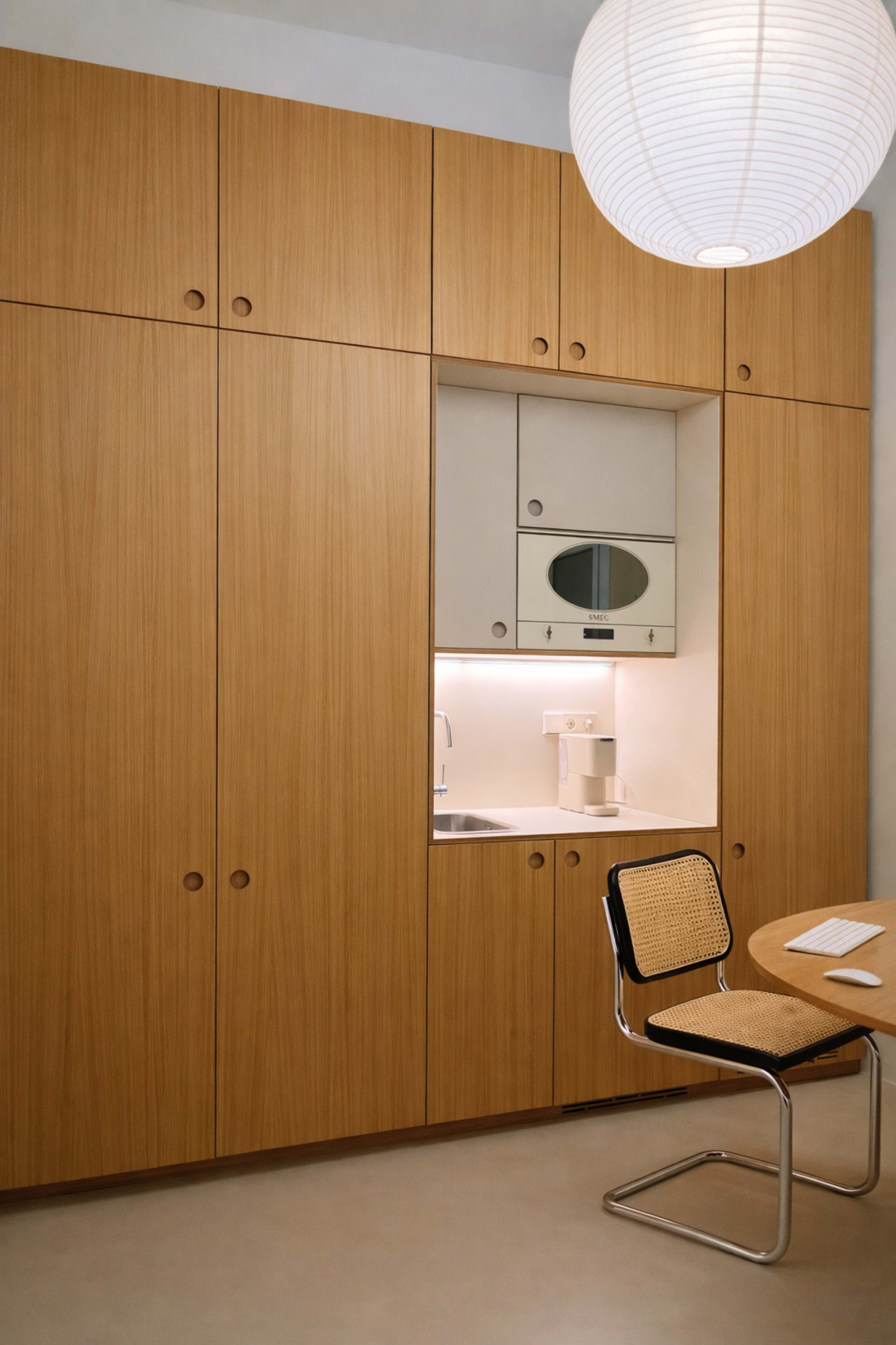 The image shows a modern kitchen with wooden cabinetry, a small counter space, and a circular pendant light overhead.