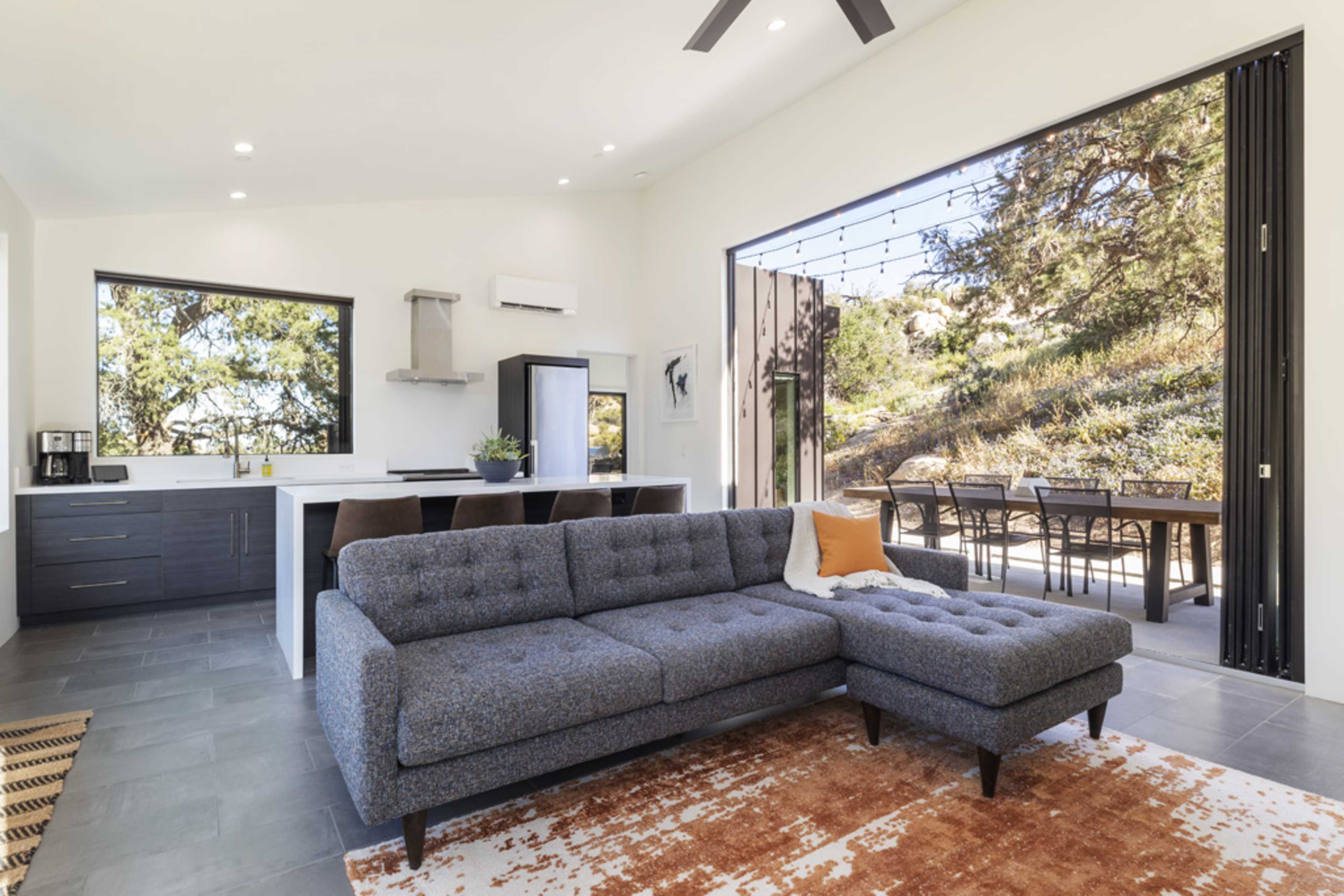 A modern living room features a gray sectional sofa facing an open space with a kitchen and large sliding doors leading to an outdoor dining area.