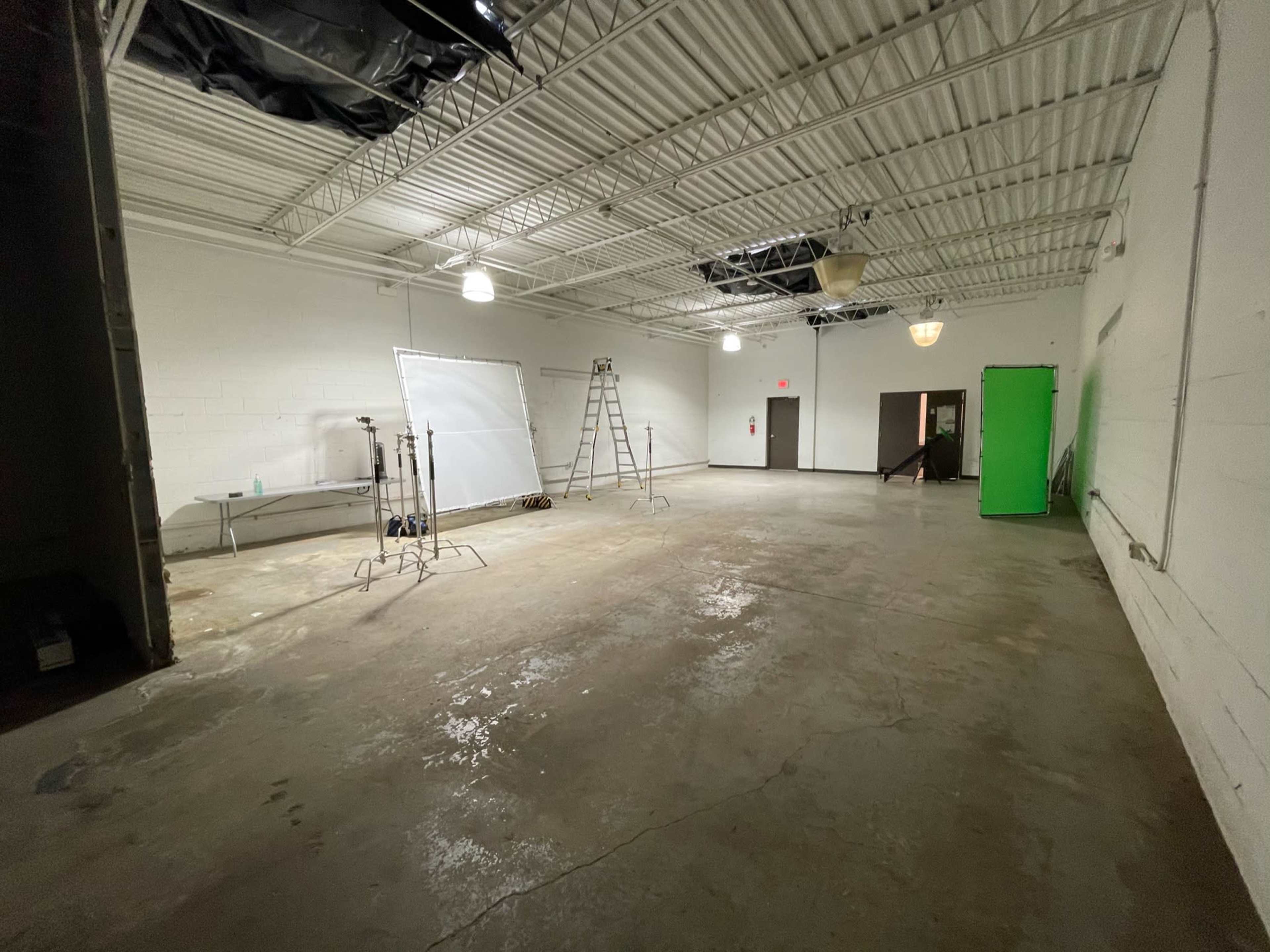 An empty industrial space with a concrete floor, a few lighting stands, a ladder, and a green backdrop set against the wall.