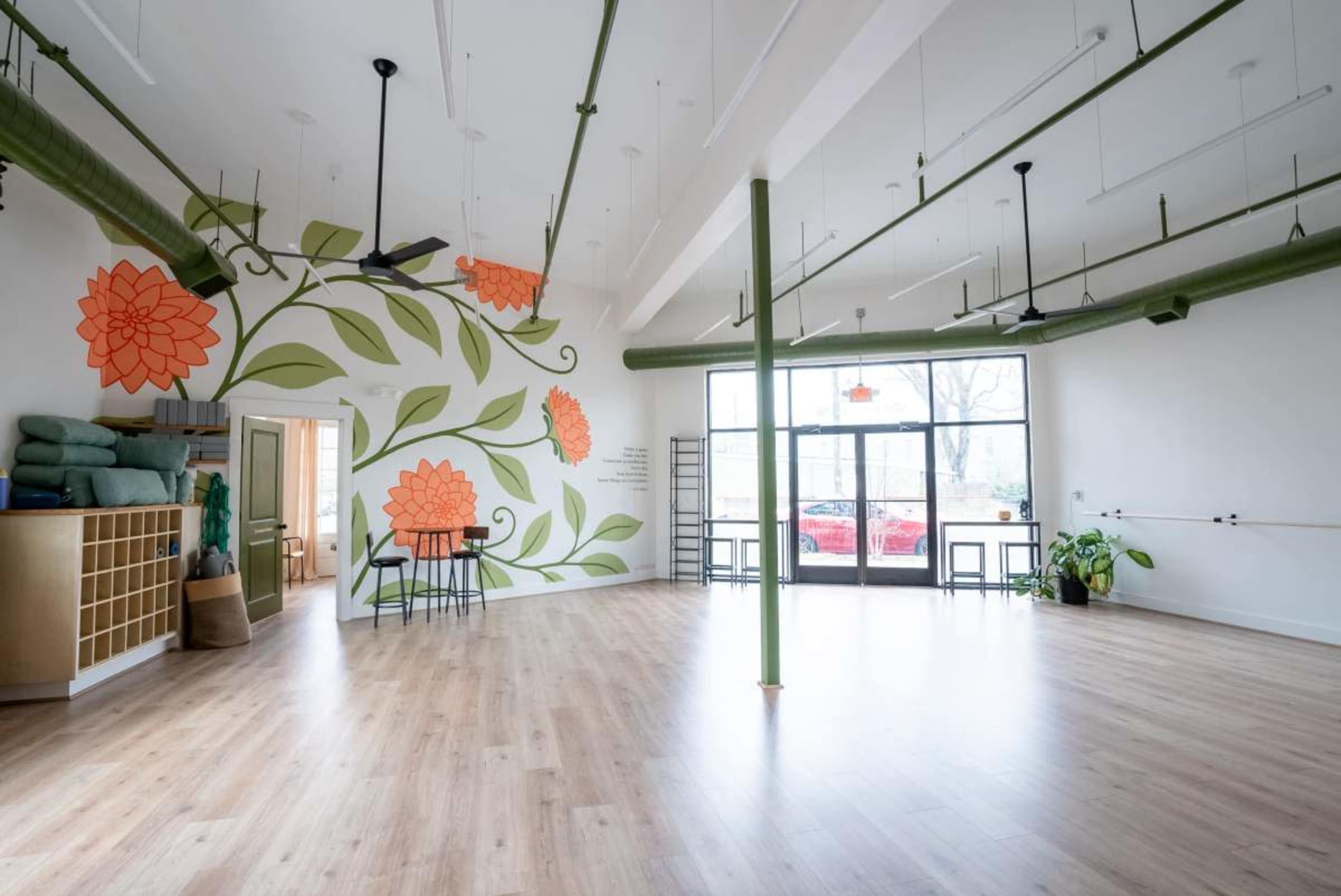 The image shows a spacious studio with a light wooden floor, large windows, and a mural of orange flowers on a white wall.