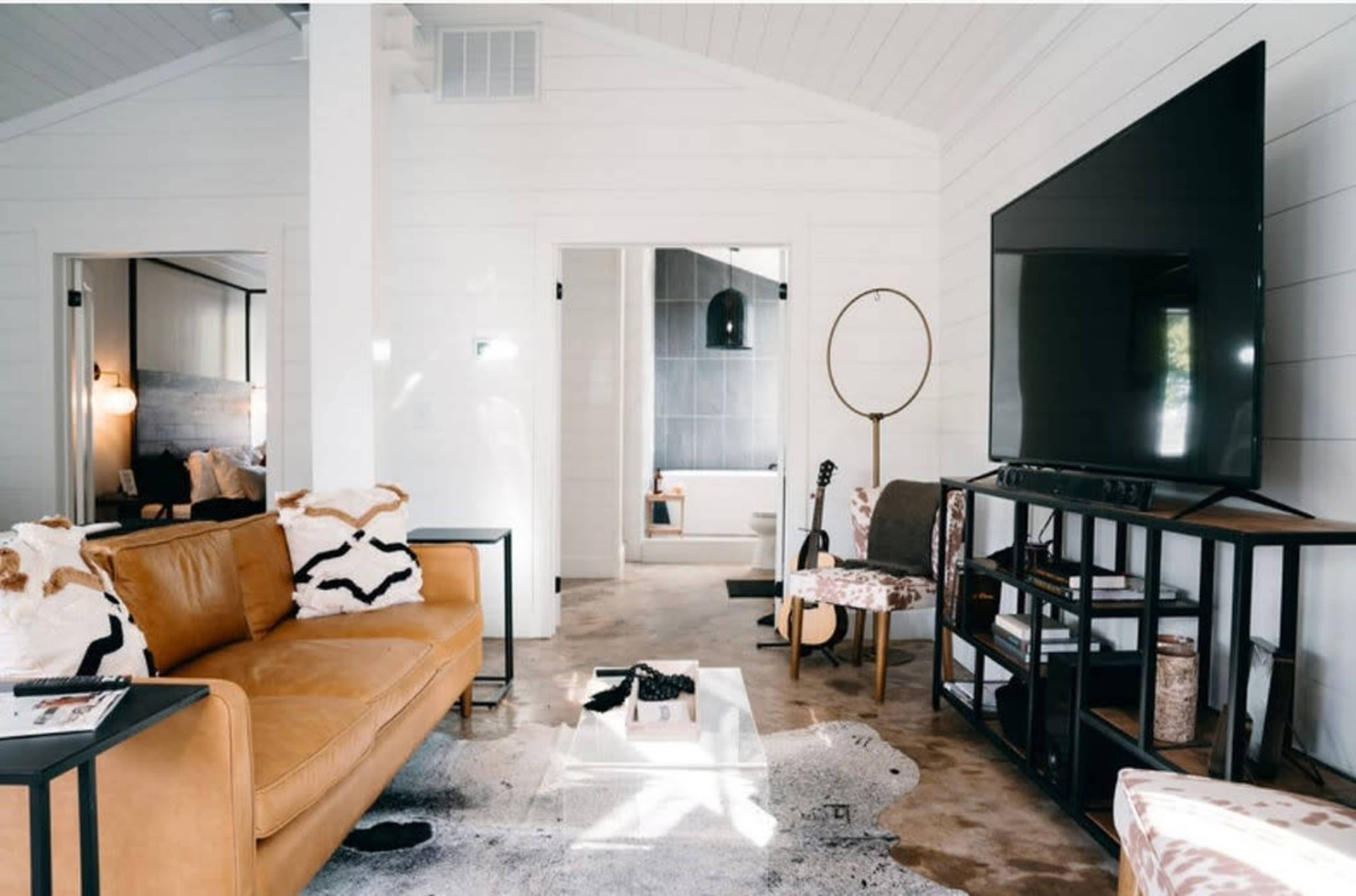 The image shows a modern living room featuring a brown leather sofa, a large television mounted on the wall, and a coffee table on a patterned rug.
