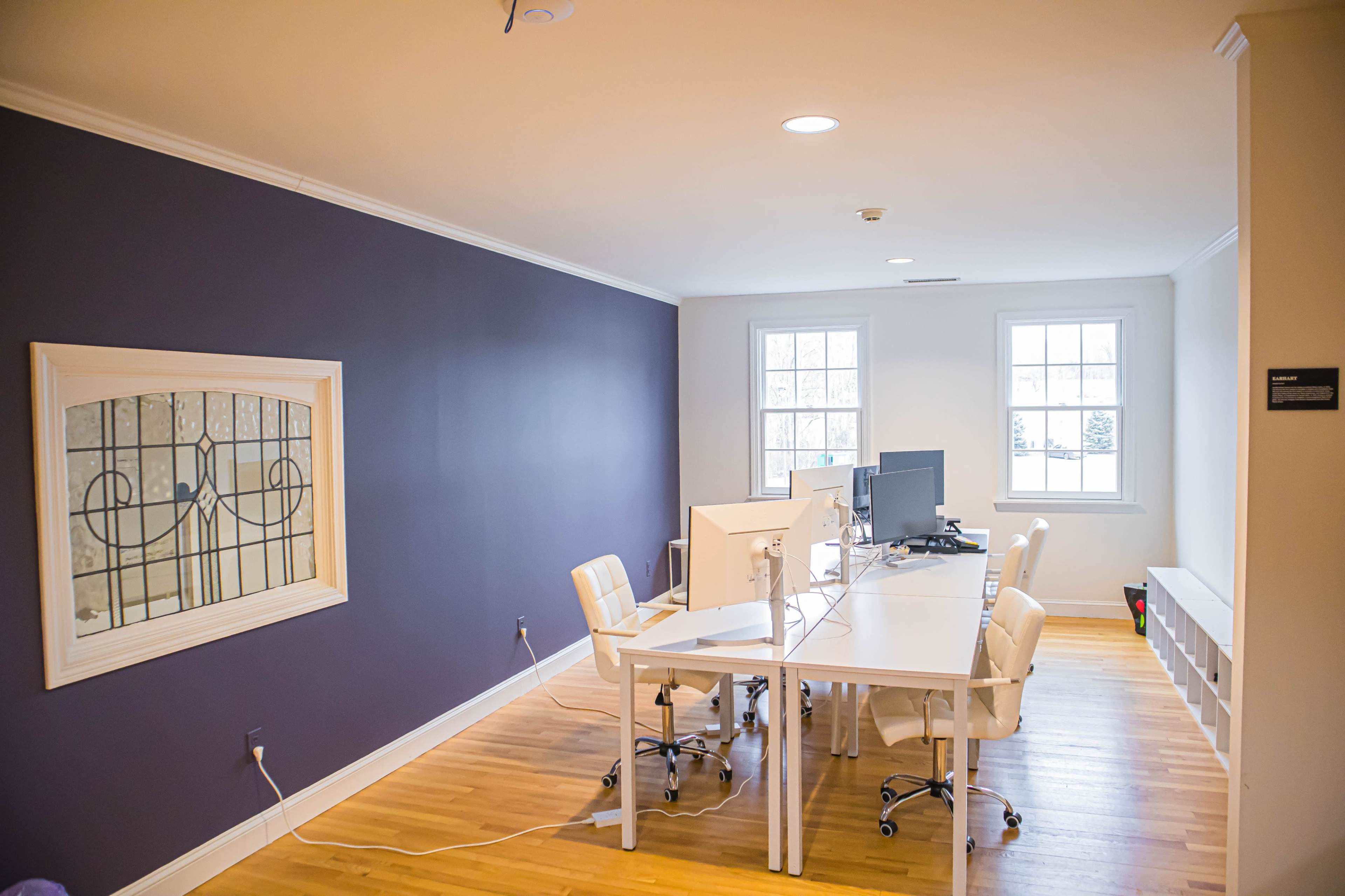A minimalist office space with a white desk, two computer monitors, and white chairs against a purple wall.