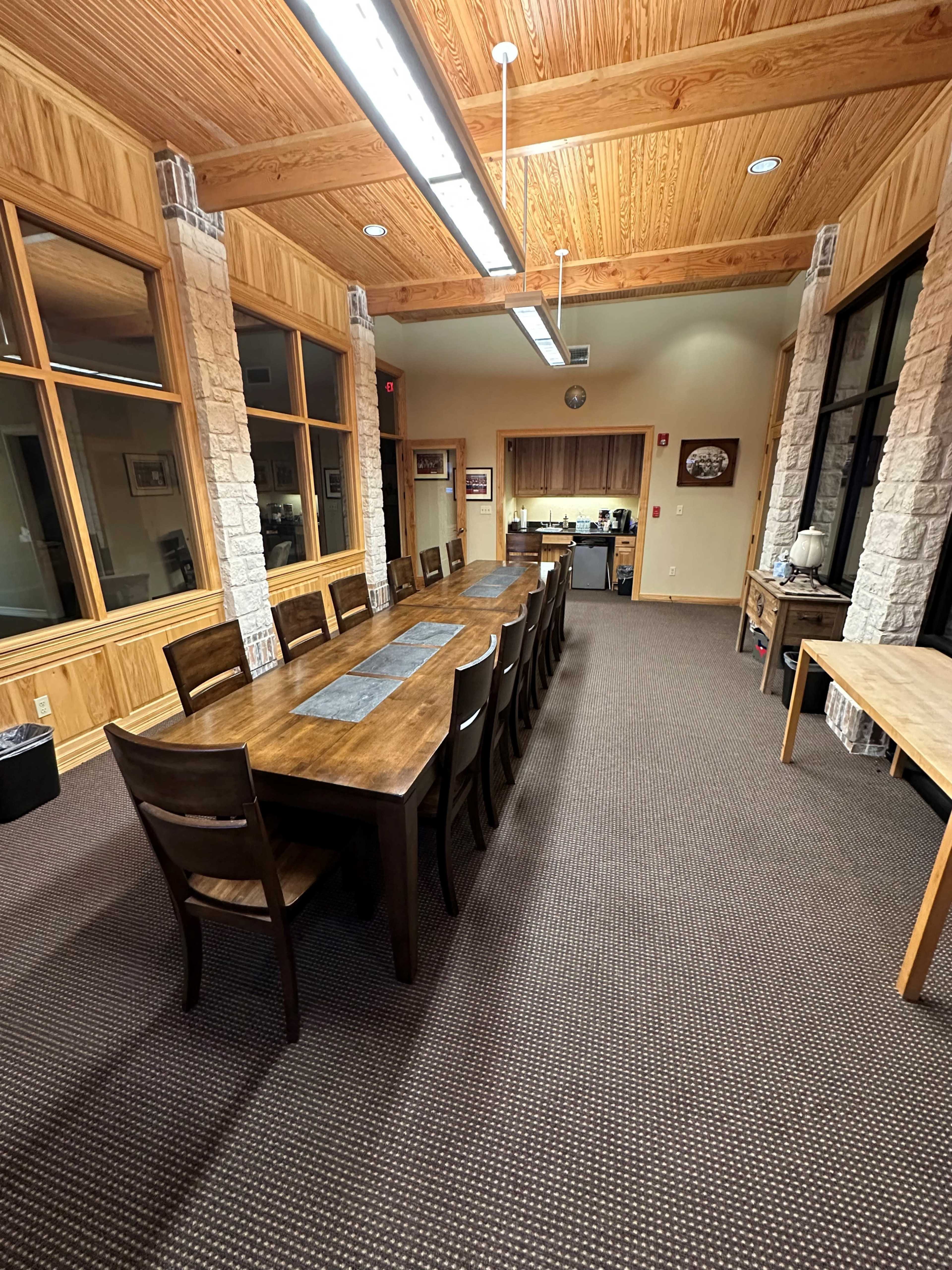 A long wooden dining table is centered in a spacious room with large windows and wooden accents.
