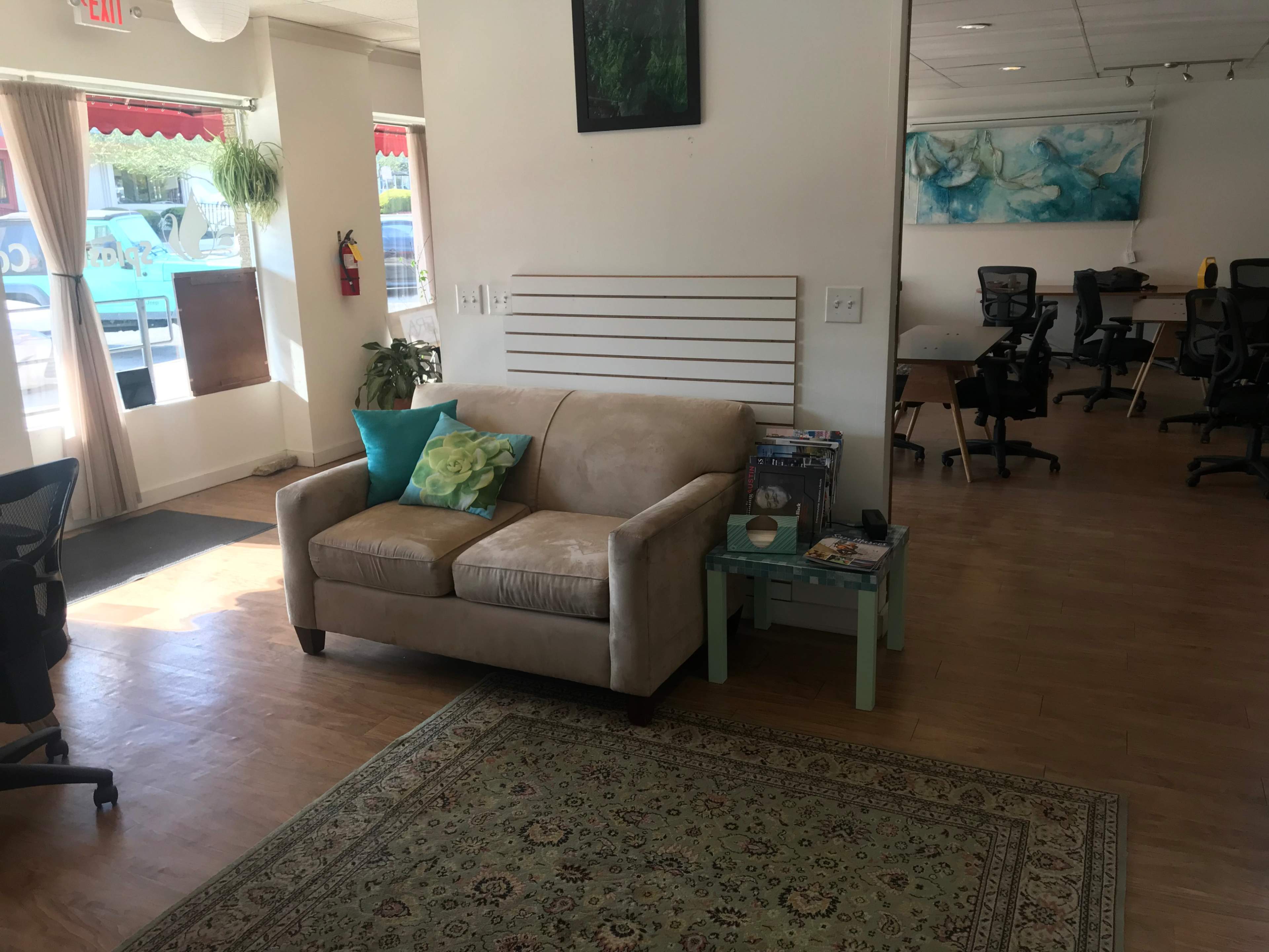 A light-filled interior with a beige sofa, a small green table, and workstations in the background.
