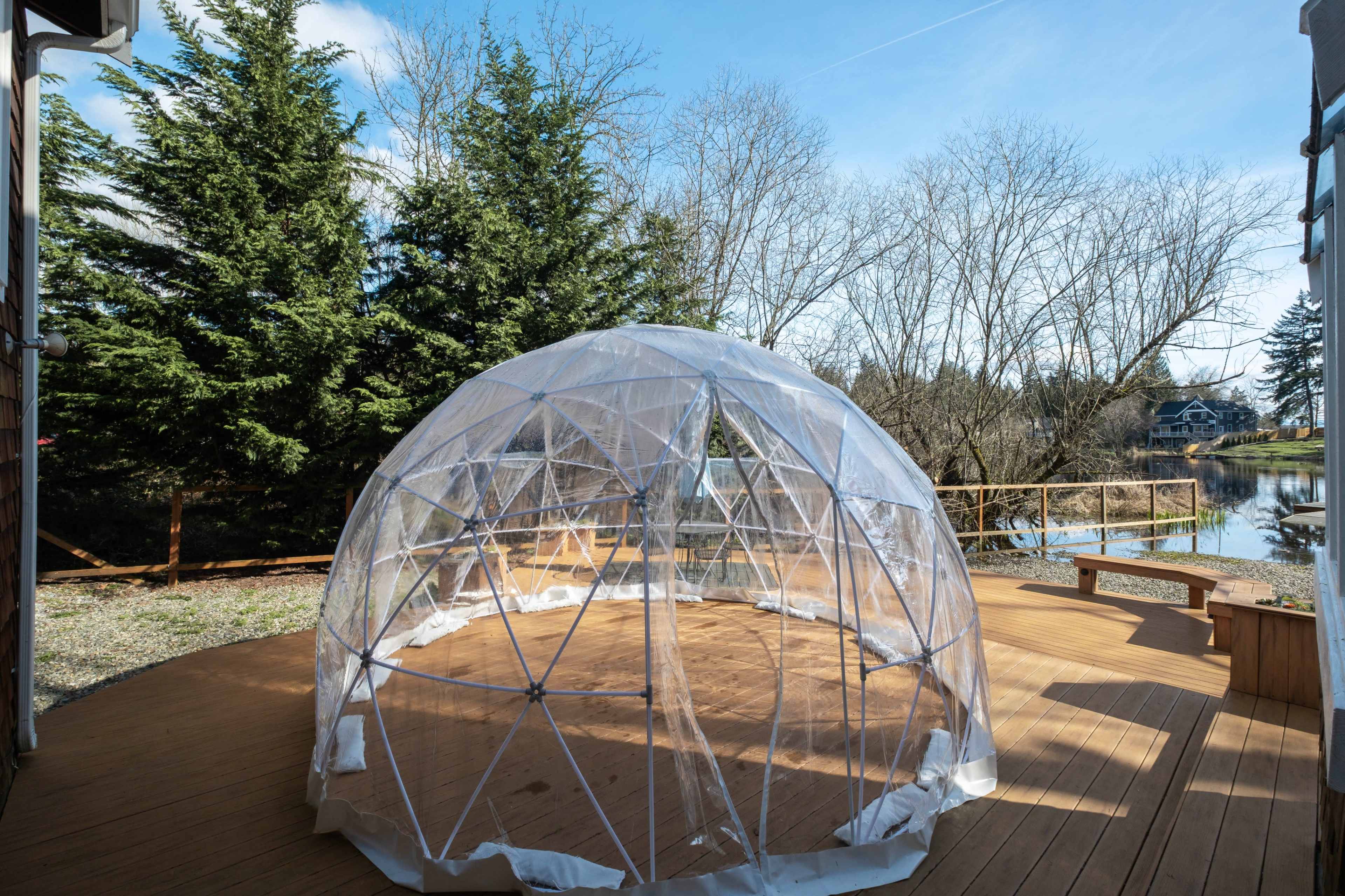 A transparent geodesic dome sits on a wooden deck surrounded by trees and a reflective body of water.