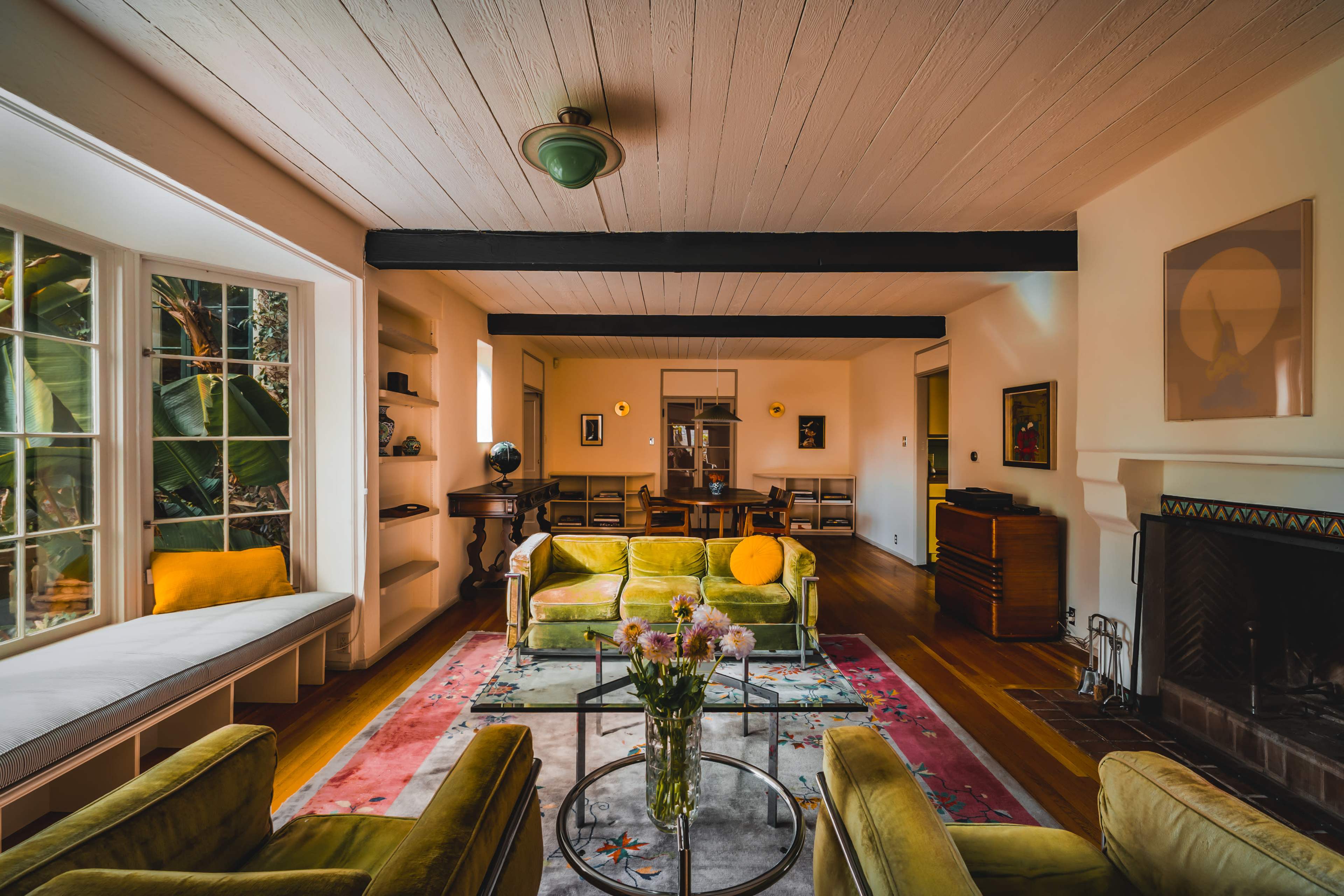 The living room features green velvet seating arranged around a glass coffee table, with large windows letting in natural light and colorful artwork adorning the walls.