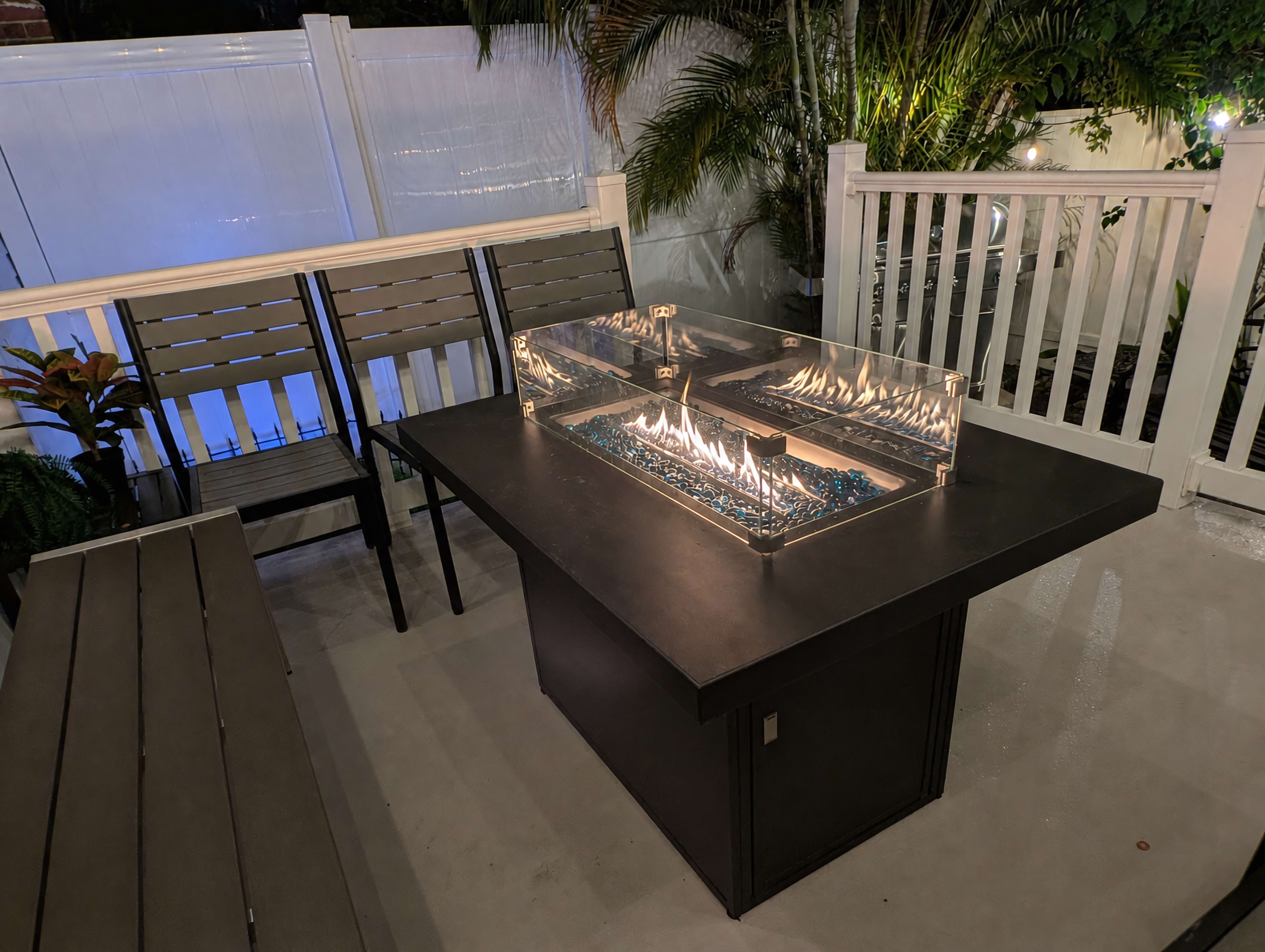 A modern outdoor dining table featuring a fire pit in the center, surrounded by blue glass stones, with seating nearby and tropical plants in the background.