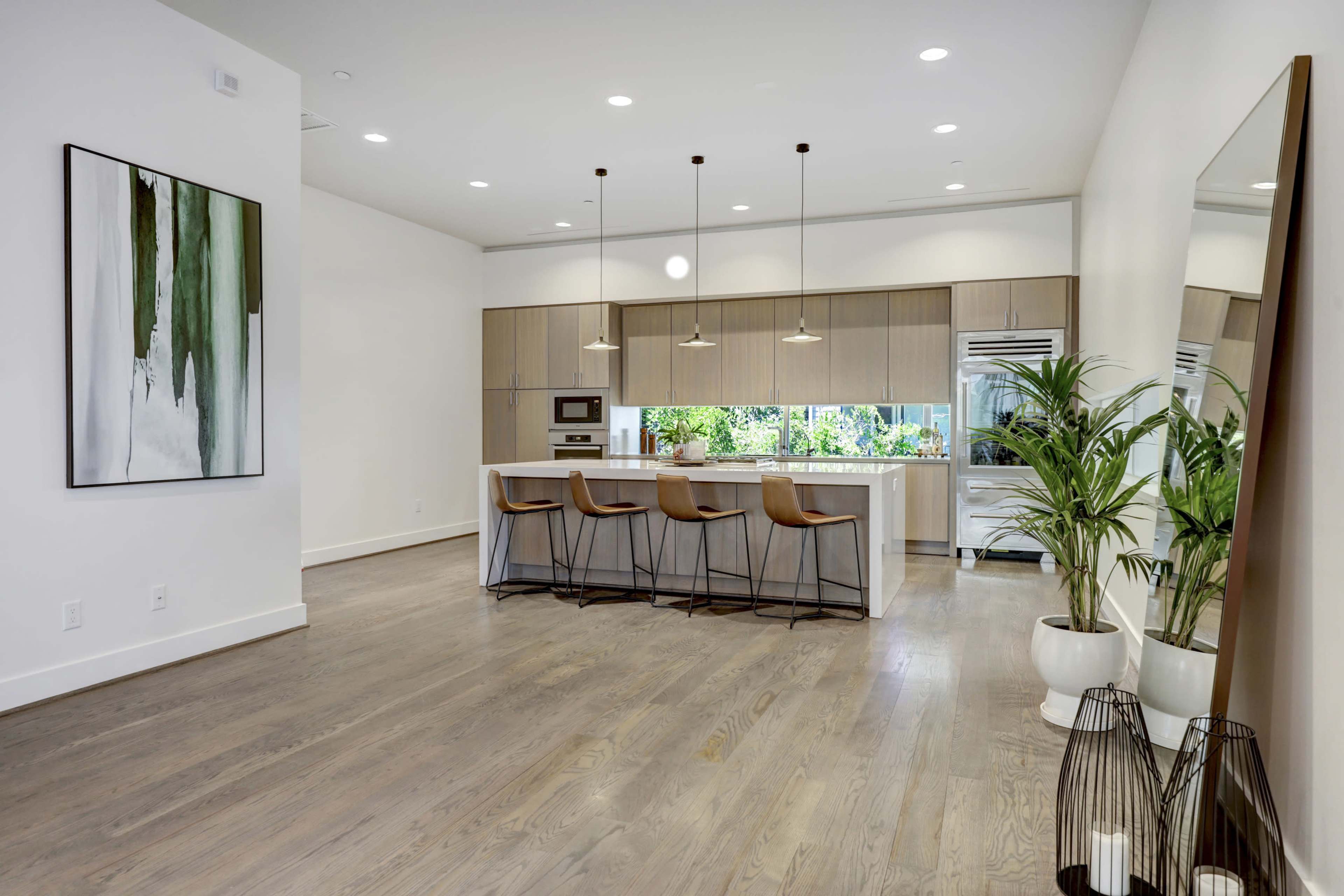 A modern kitchen features a central island with three pendant lights, sleek cabinetry, and large windows overlooking greenery.