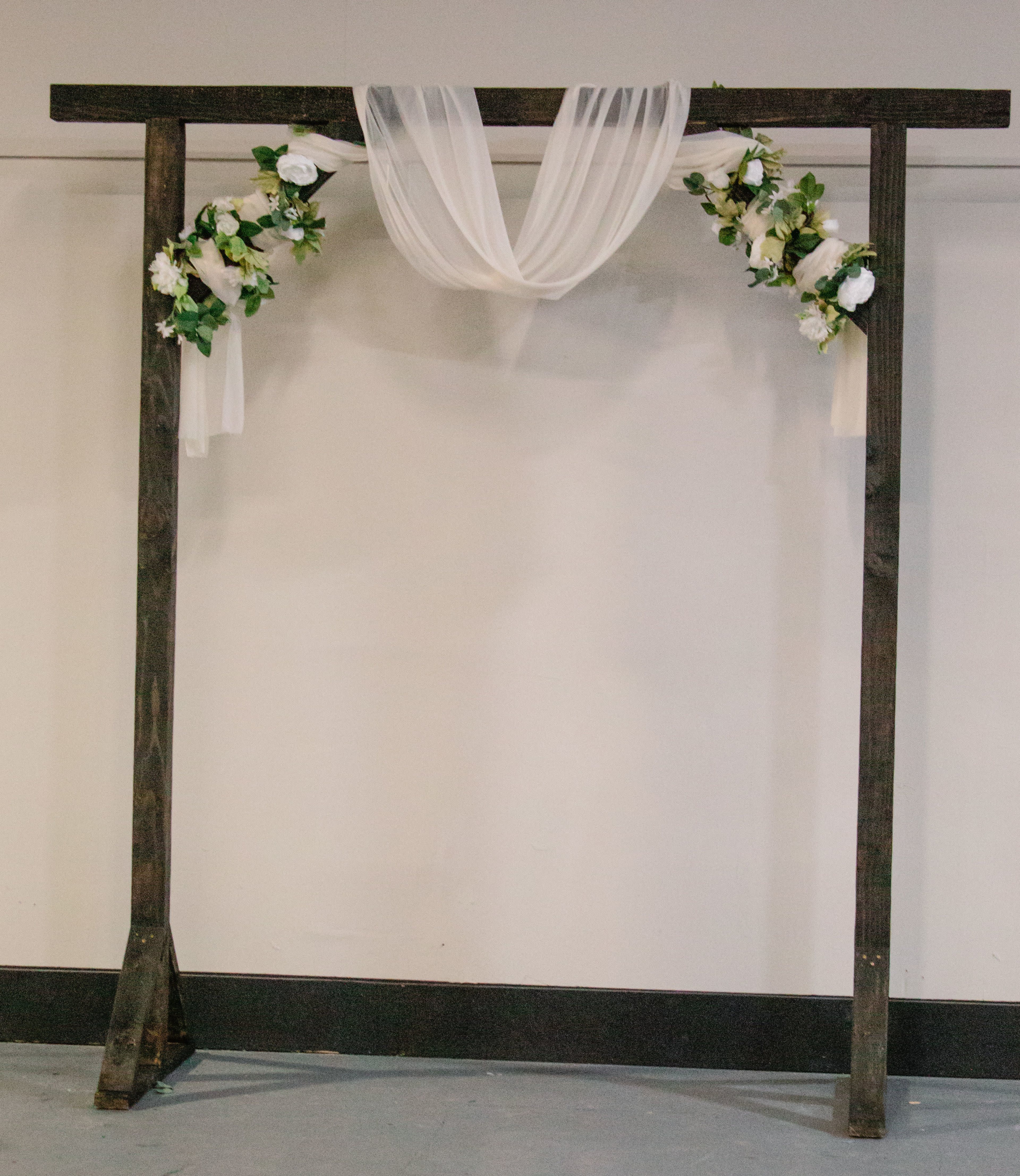 A wooden arch draped with sheer fabric and adorned with white flowers is positioned against a plain wall.