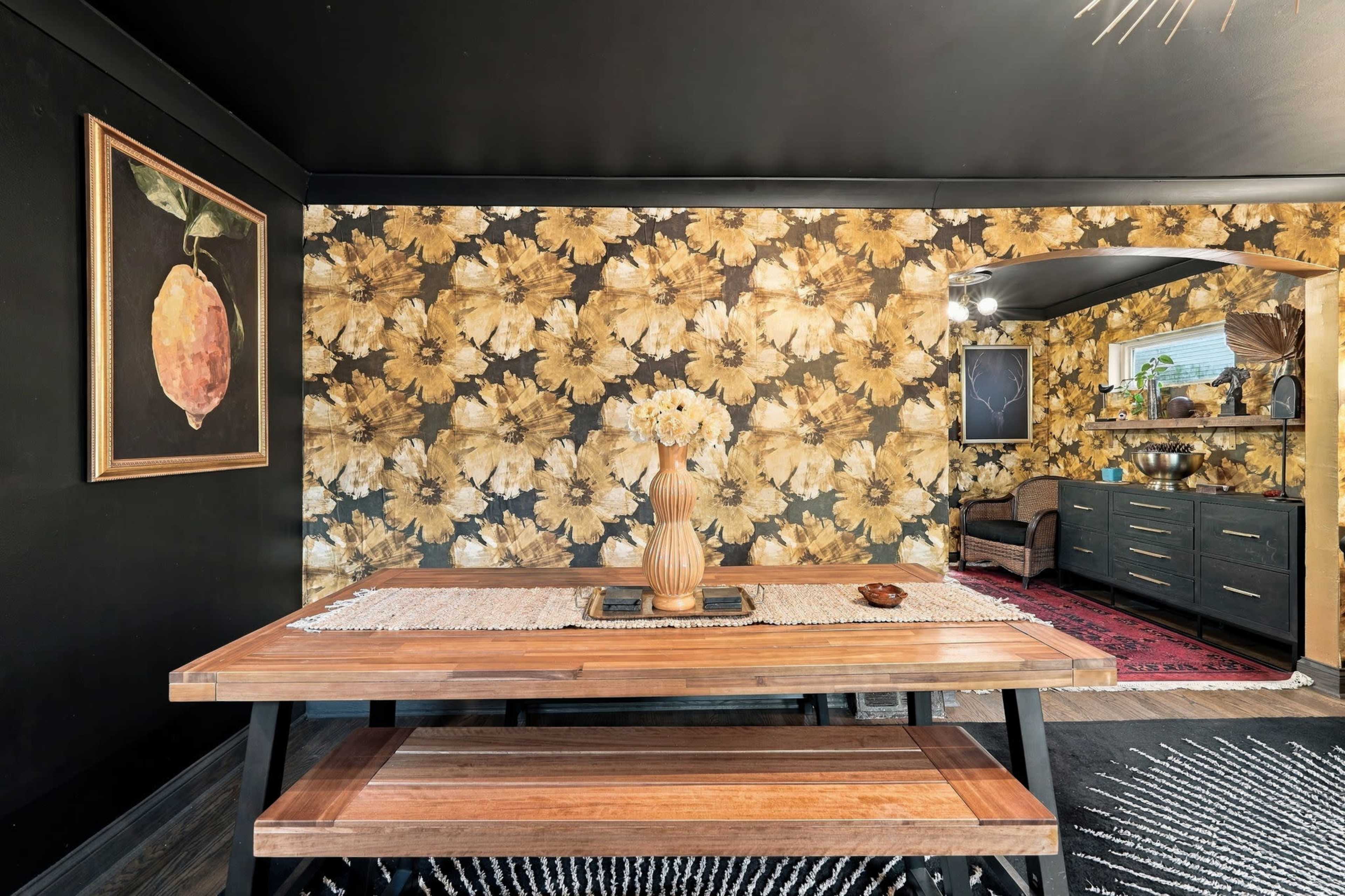 A modern dining area features a wooden table with a vase of flowers, surrounded by floral wallpaper and dark accents.