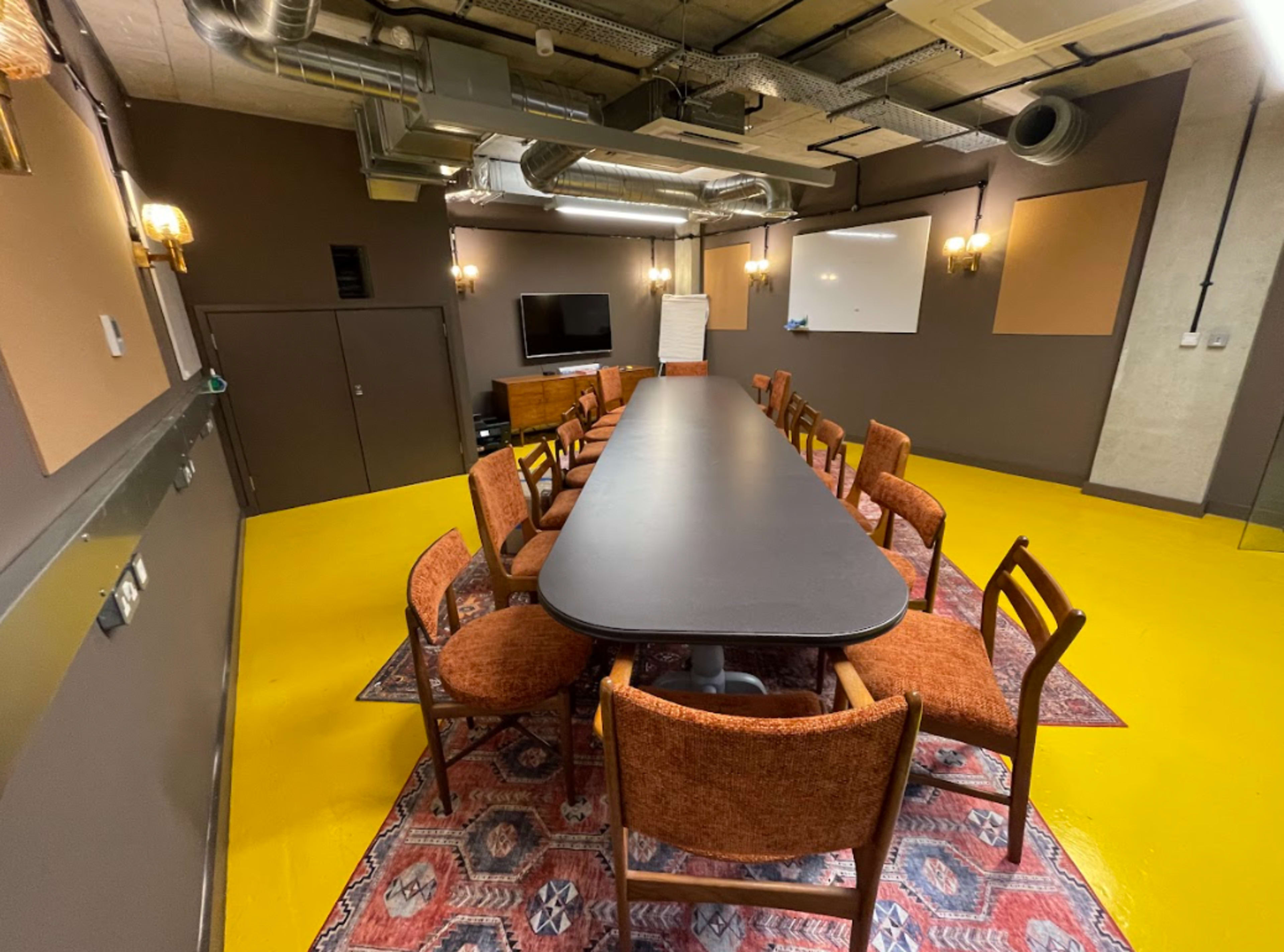 A conference room features a long black table surrounded by wooden chairs on a yellow floor, with a television and whiteboard mounted on the walls.