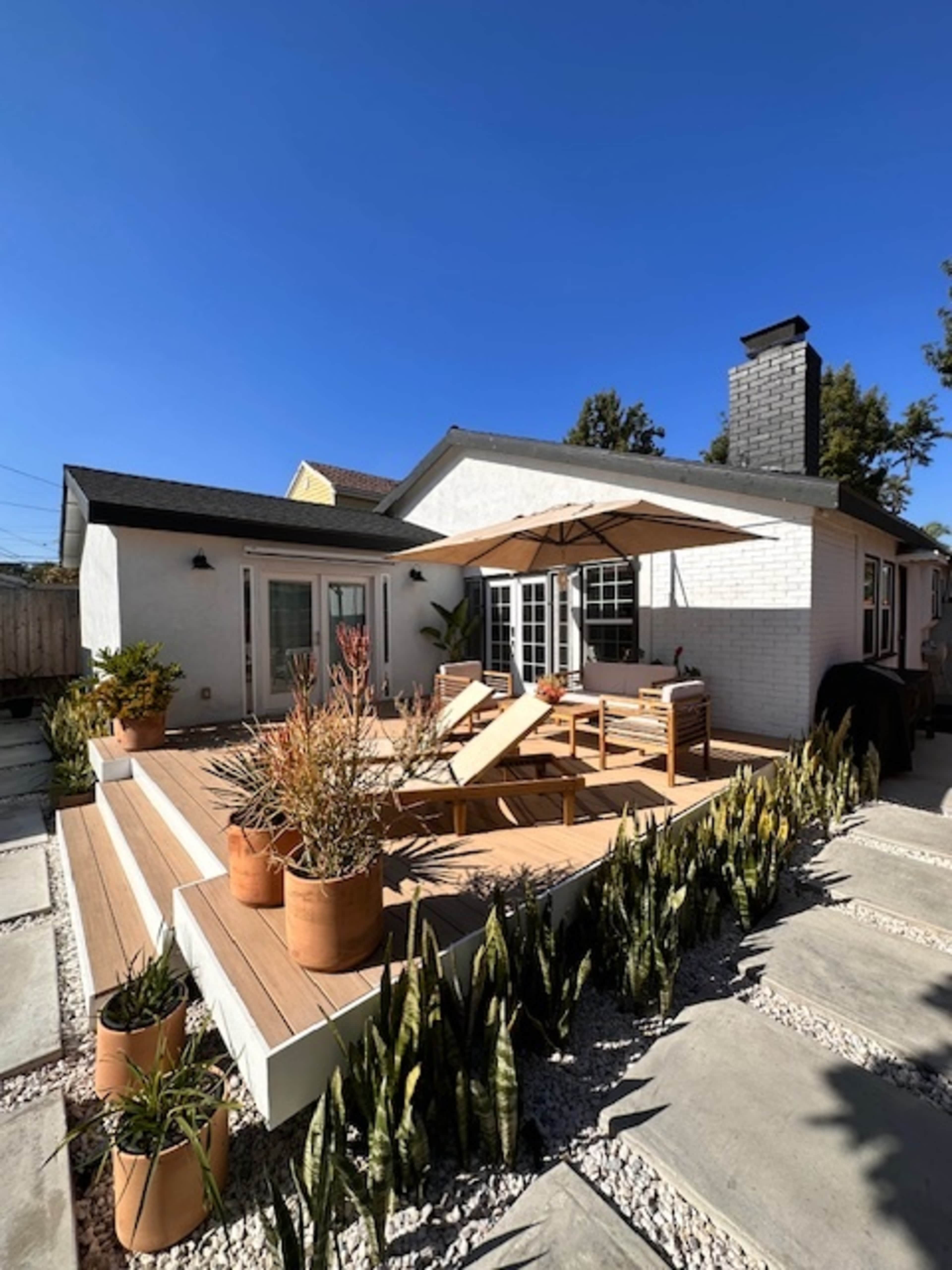 A modern home features a spacious wooden deck with lounge chairs, an umbrella, and potted plants under a clear blue sky.