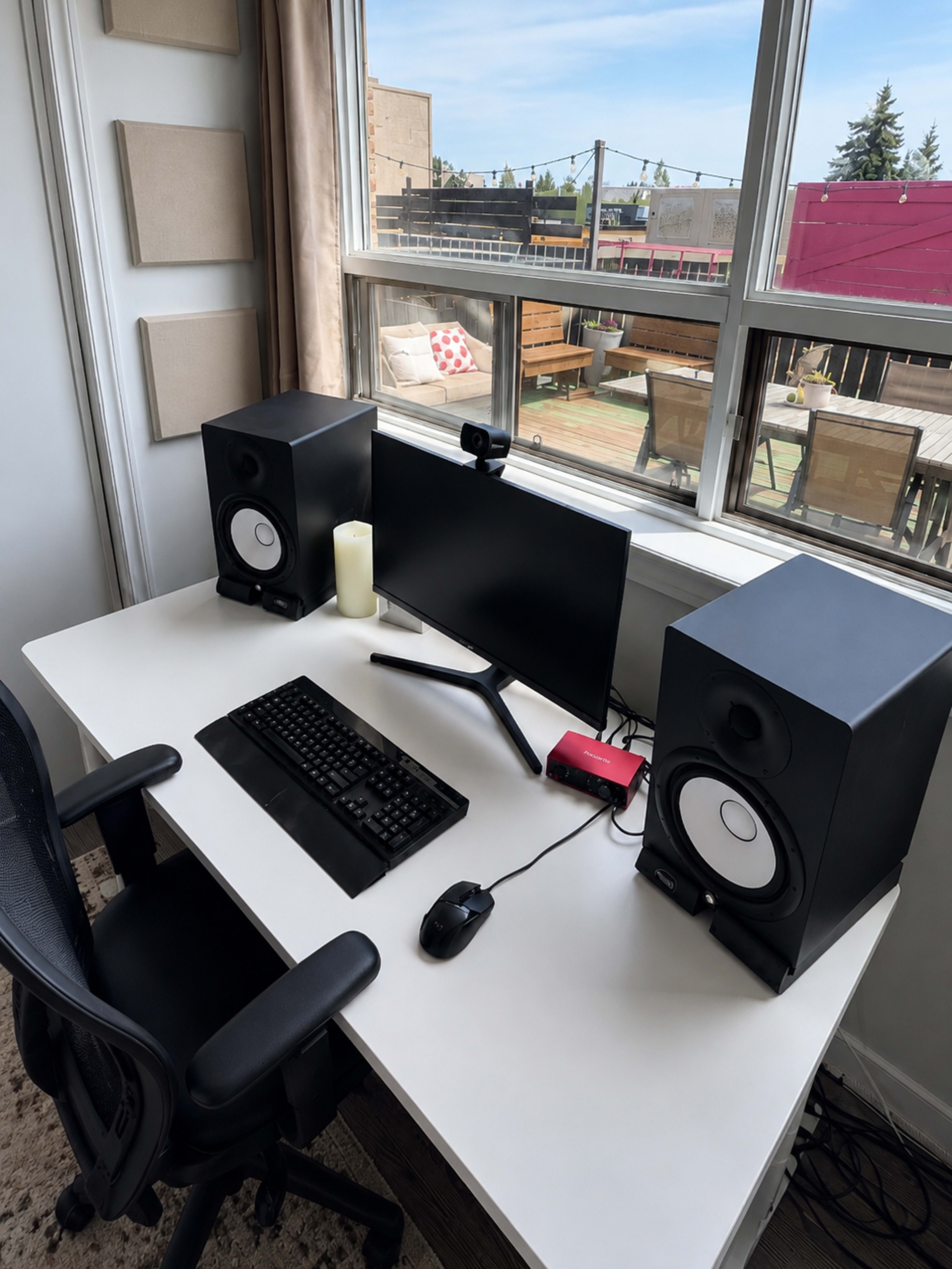 A desk setup features a computer monitor, keyboard, mouse, and two speakers, with a view of an outdoor area through large windows.