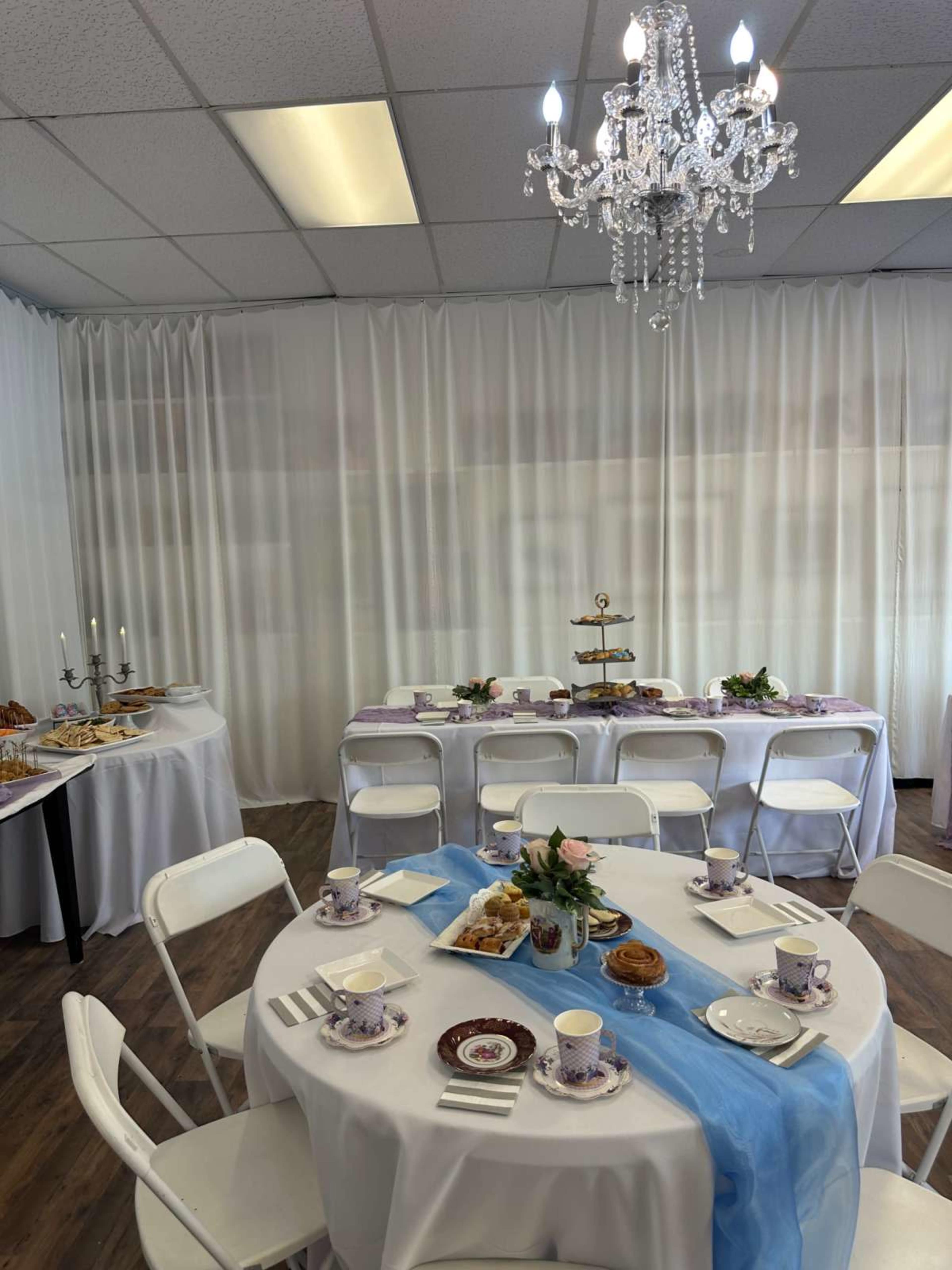 A brightly lit room features a table set for tea with floral dishes, a blue tablecloth, and a tiered serving stand, while a smaller table displays assorted pastries.
