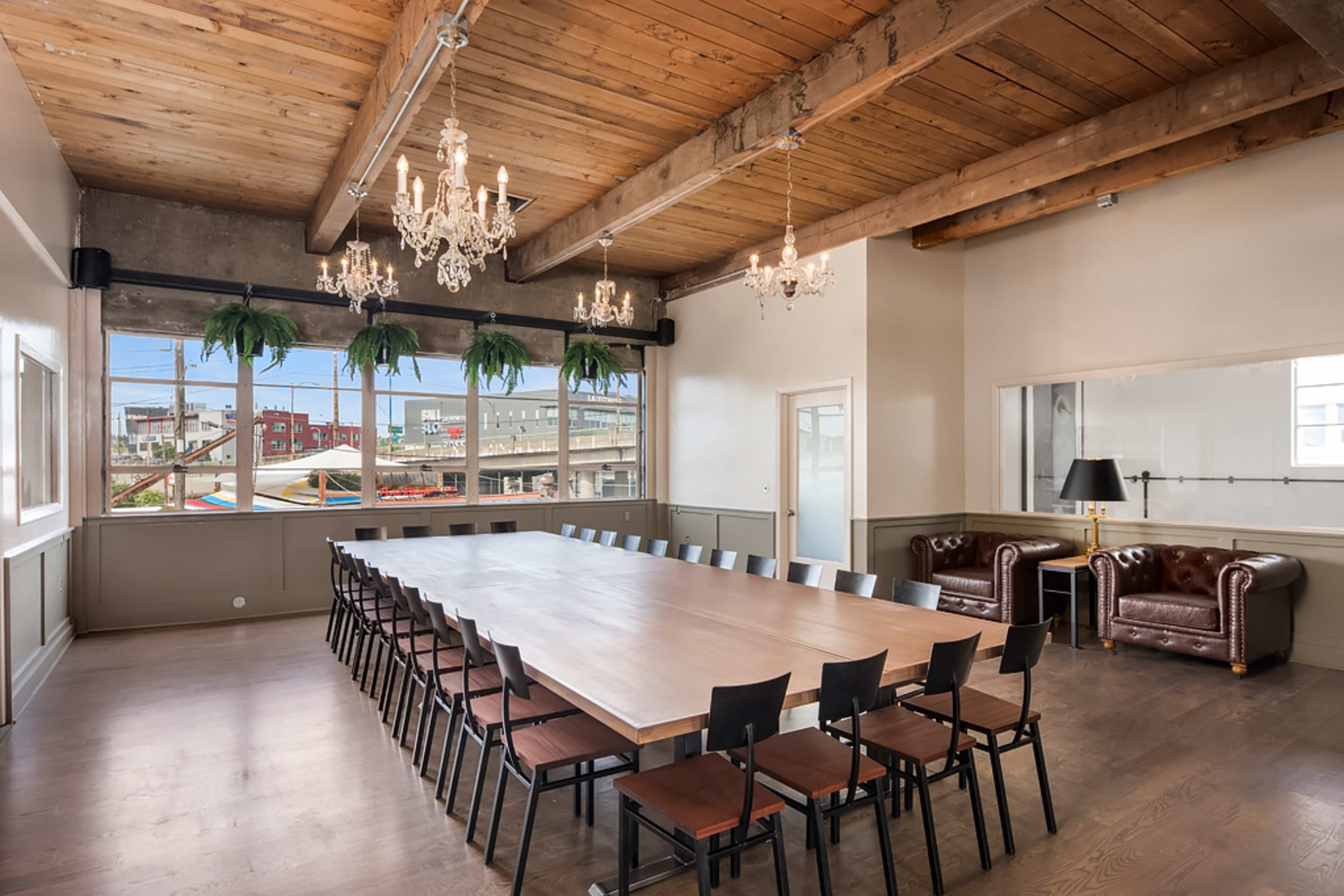 A long wooden conference table is surrounded by black chairs in a well-lit meeting room featuring exposed wooden beams and two chandeliers.