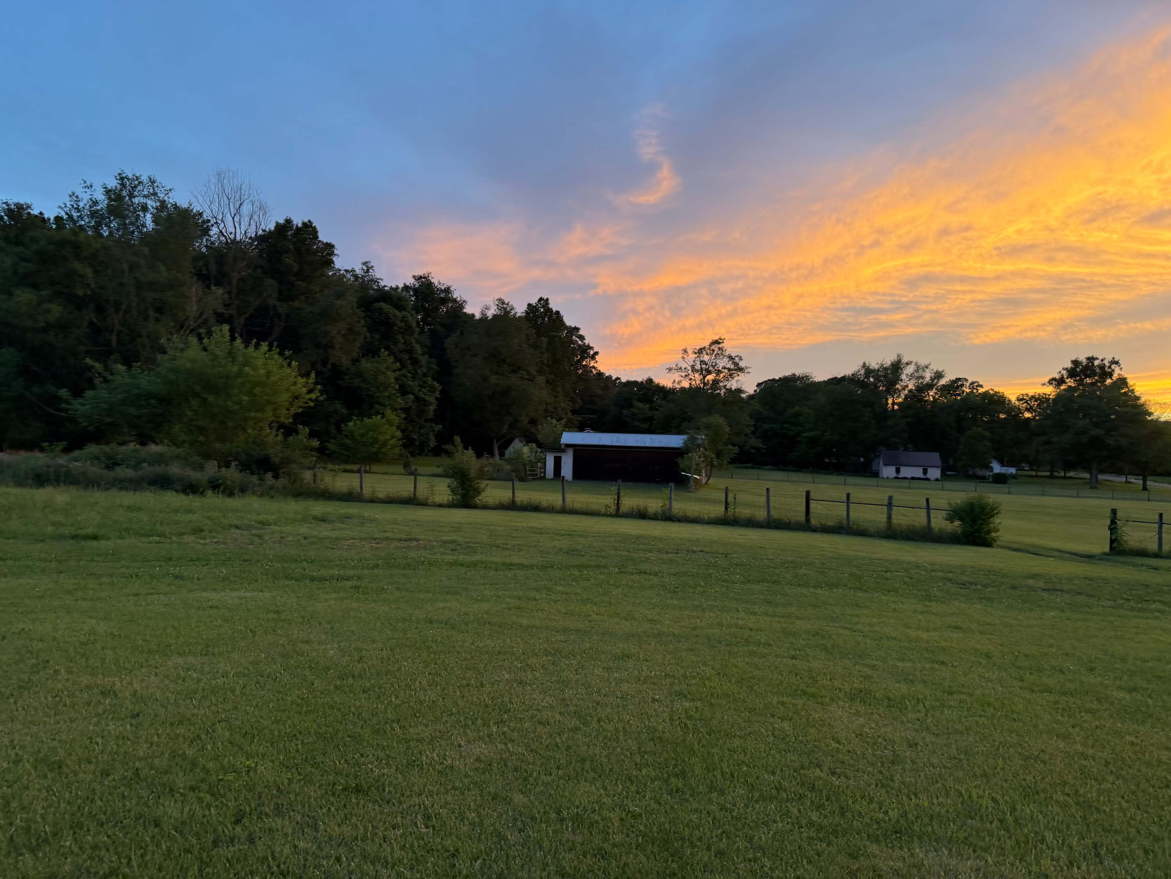 A vibrant sunset illuminates the sky above a grassy field, with two small structures nestled among the trees in the background.
