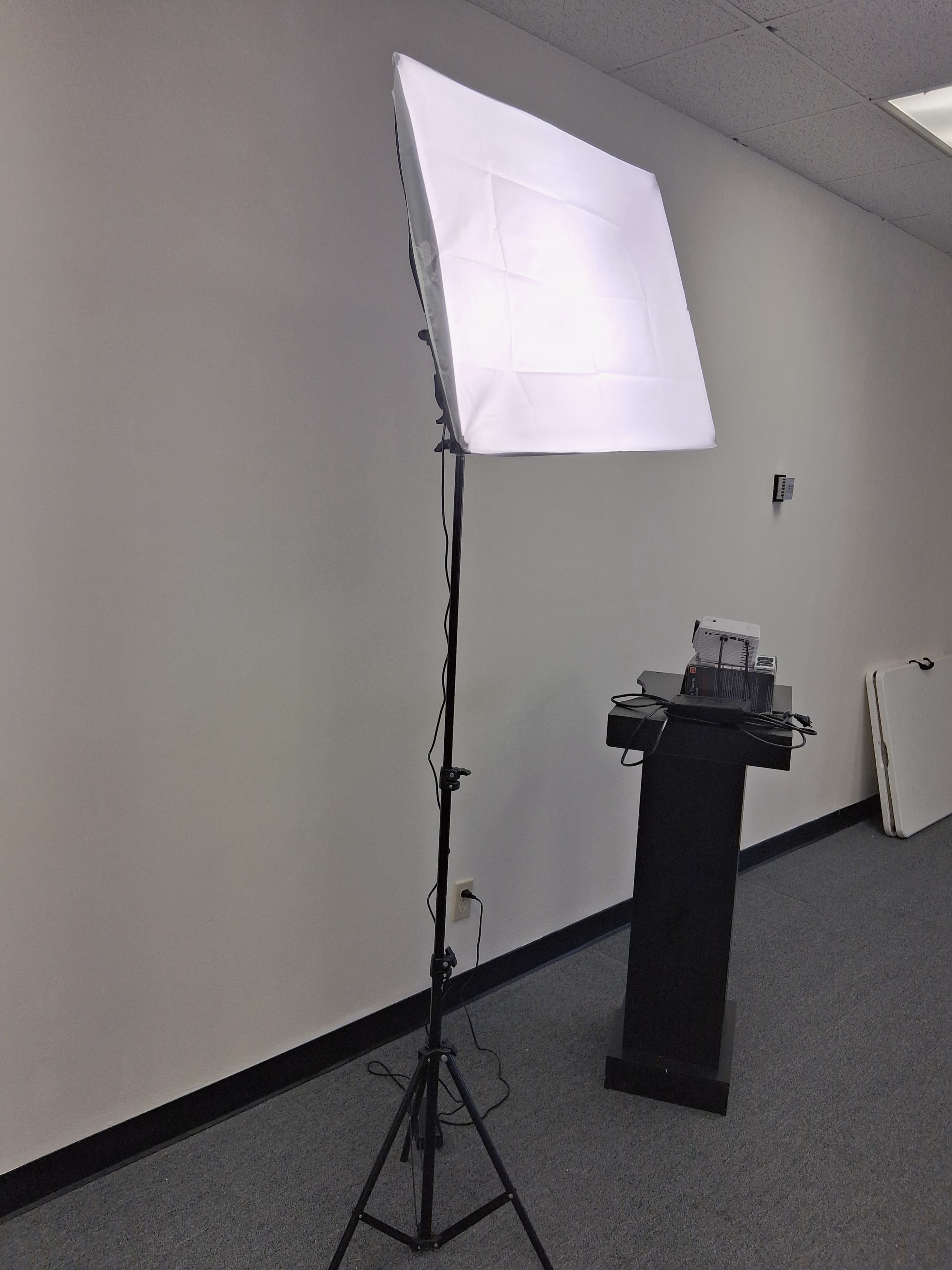 A softbox lighting setup is positioned on a stand in a room with gray carpeting and a plain wall.