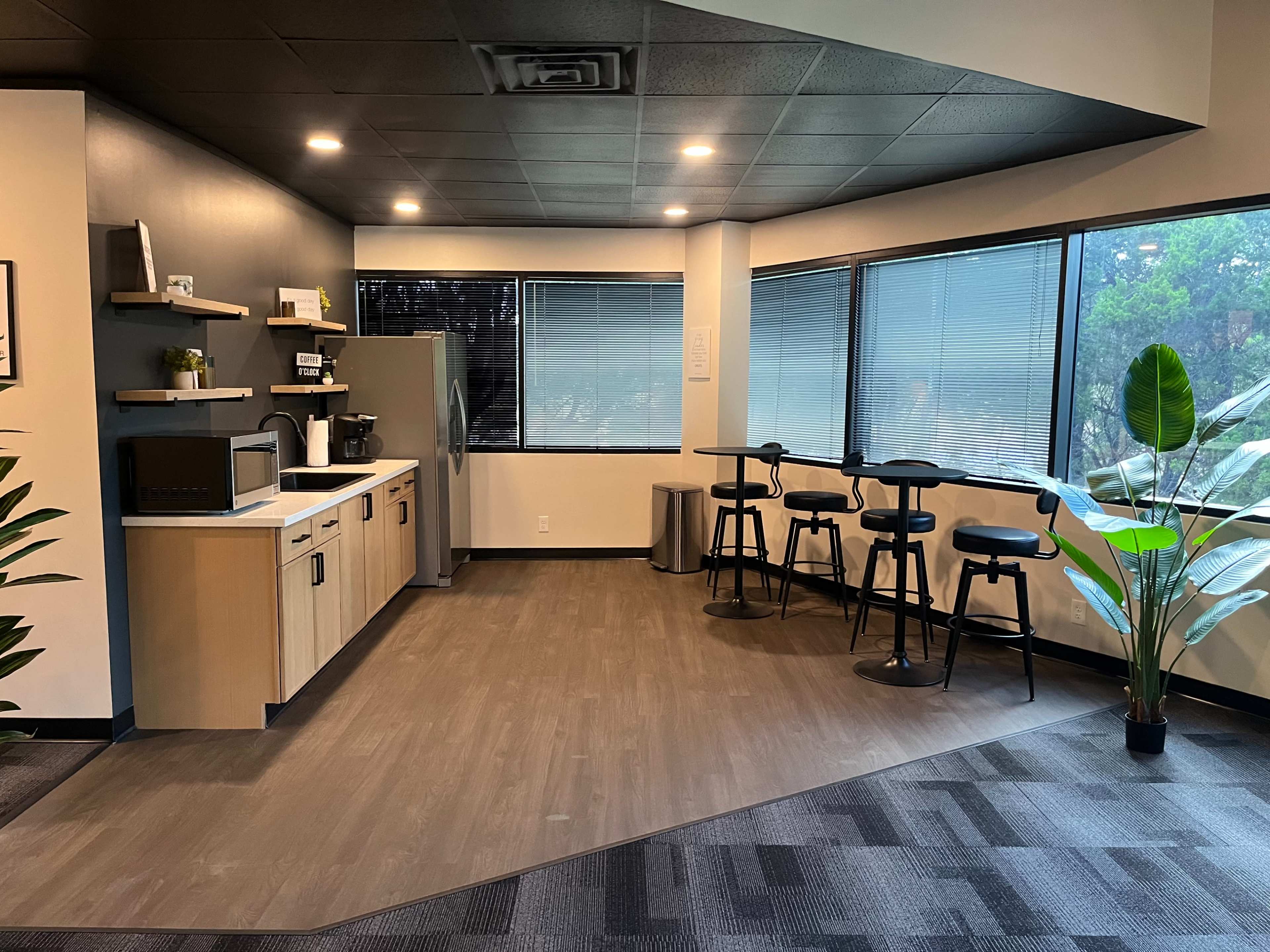 The image shows a modern break room with a kitchenette, including a refrigerator, microwave, and coffee machine, along with barstool seating and large windows.
