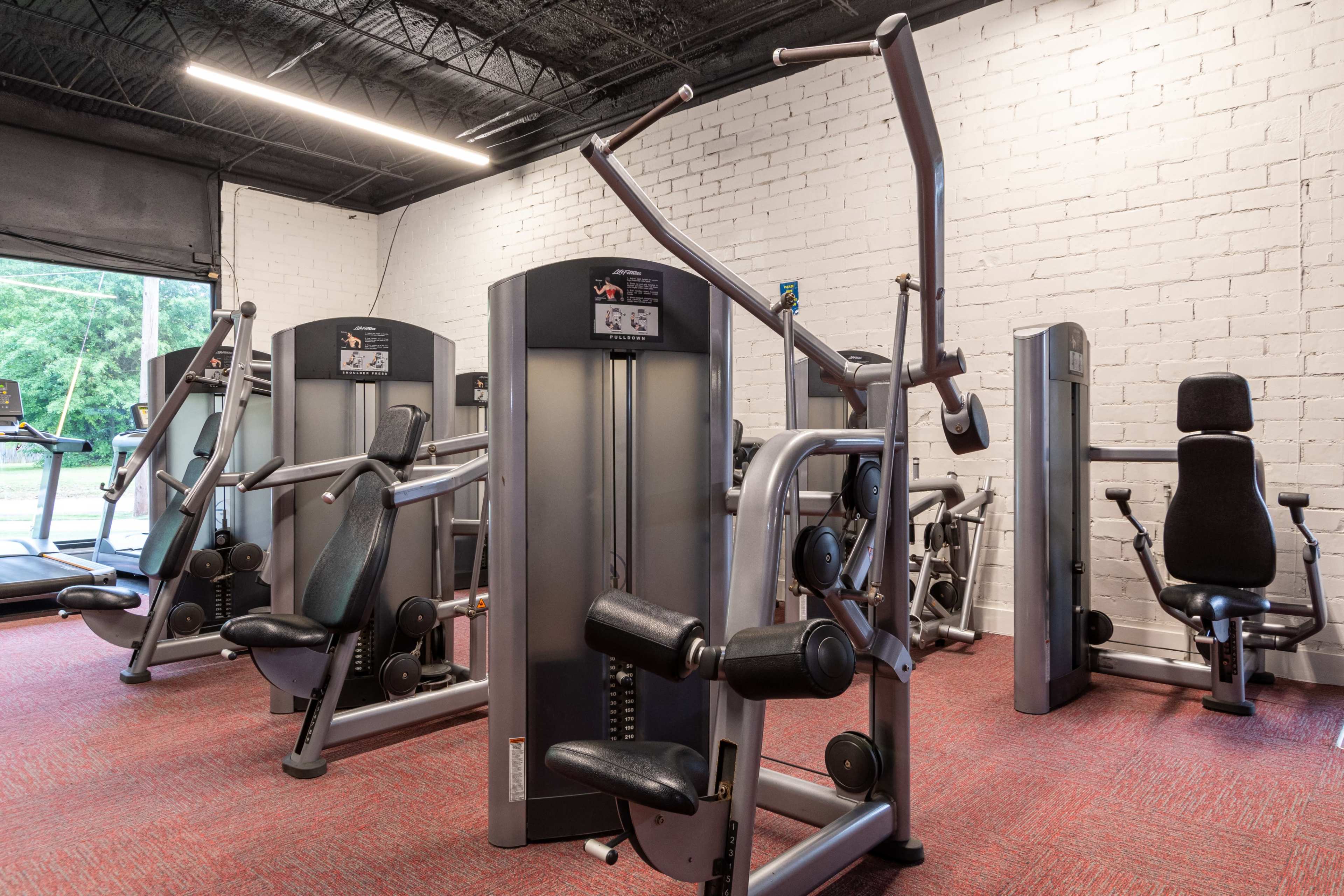 A gym interior with various weight machines arranged on a red carpeted floor against a white brick wall.
