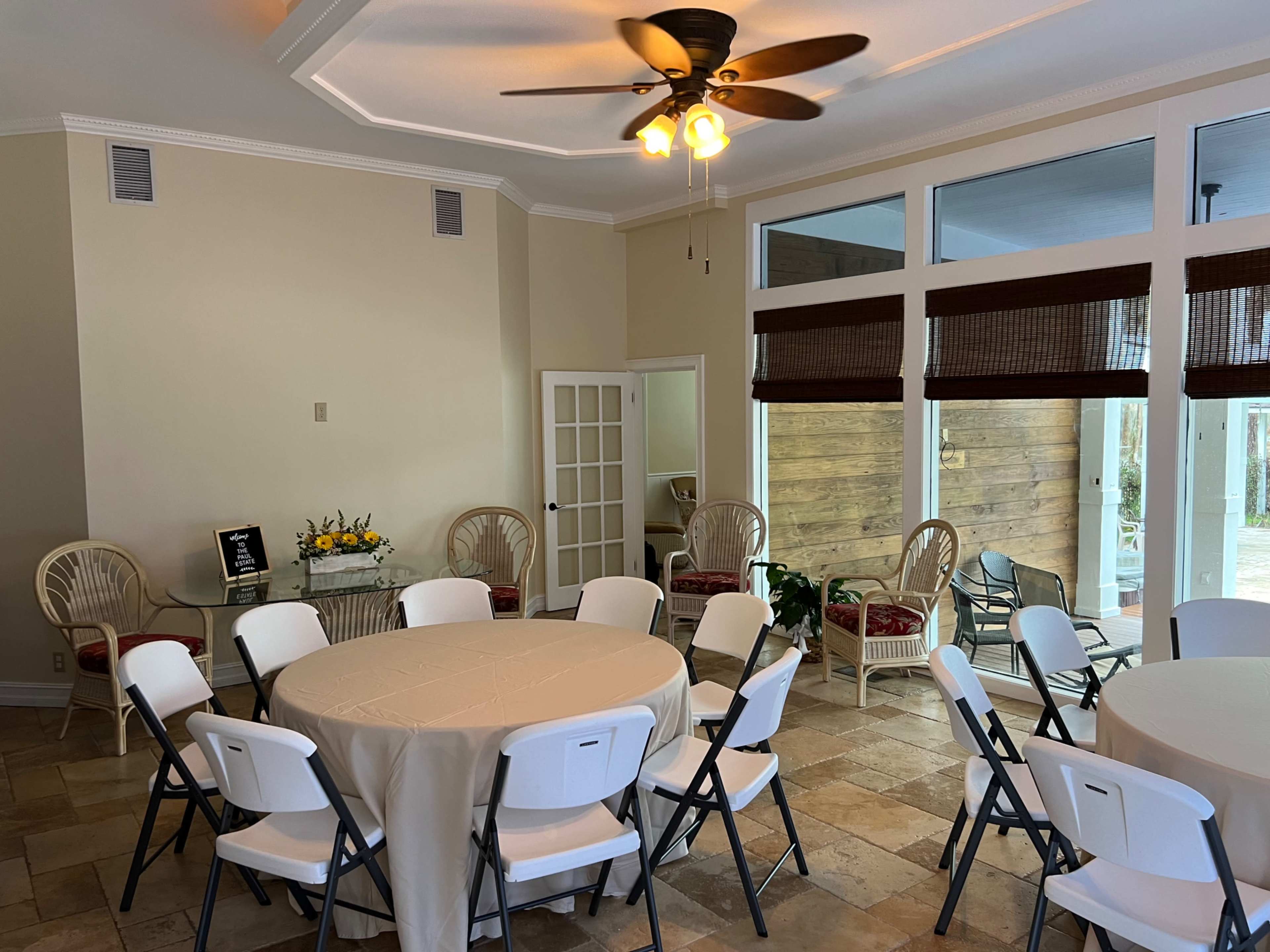 The image shows a spacious room set up with circular tables covered in white tablecloths, surrounded by folding chairs, with large windows and a small seating area in the background.