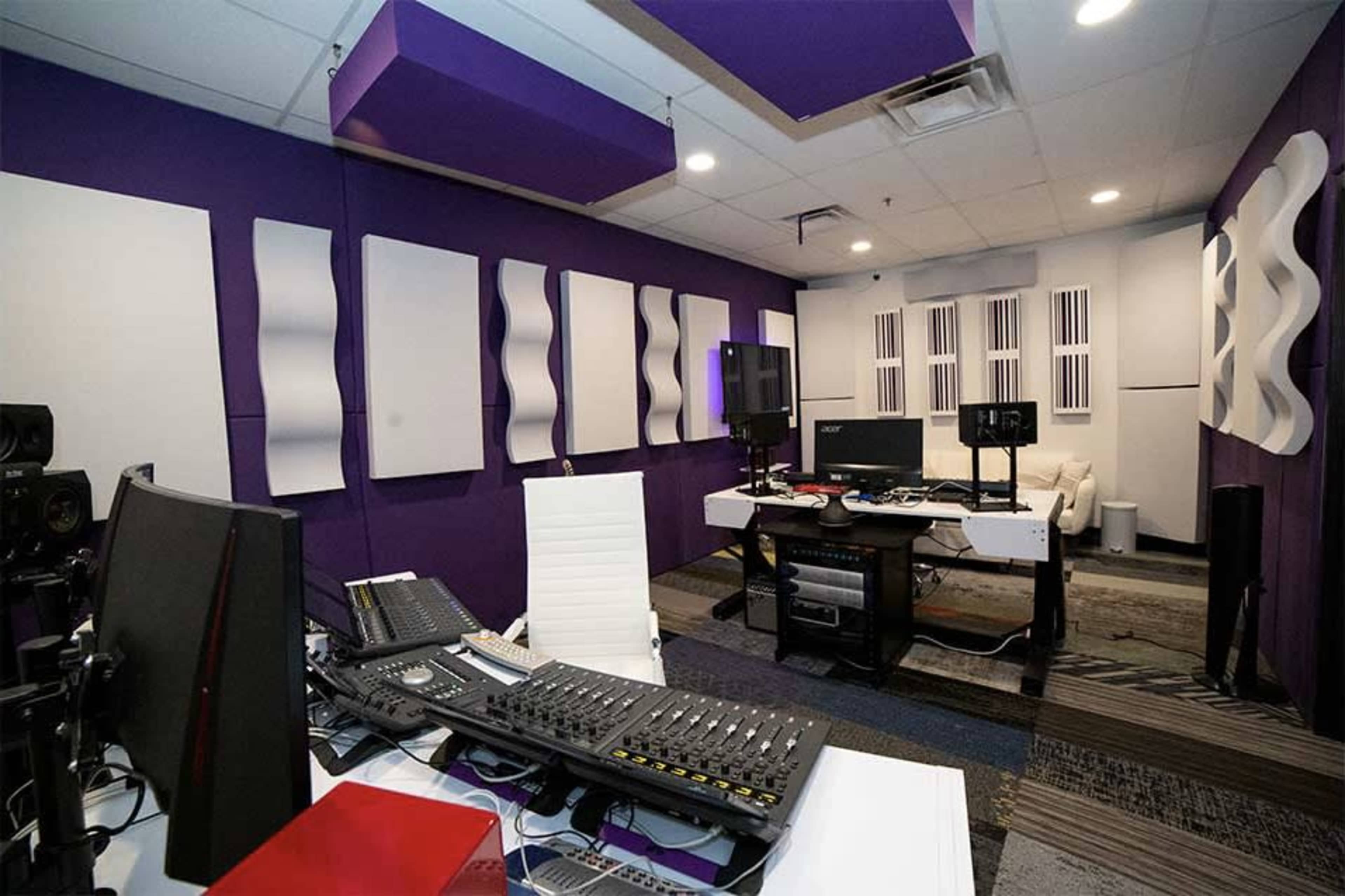 A sound recording studio with purple walls, acoustic panels, and a desk equipped with audio equipment and monitors.