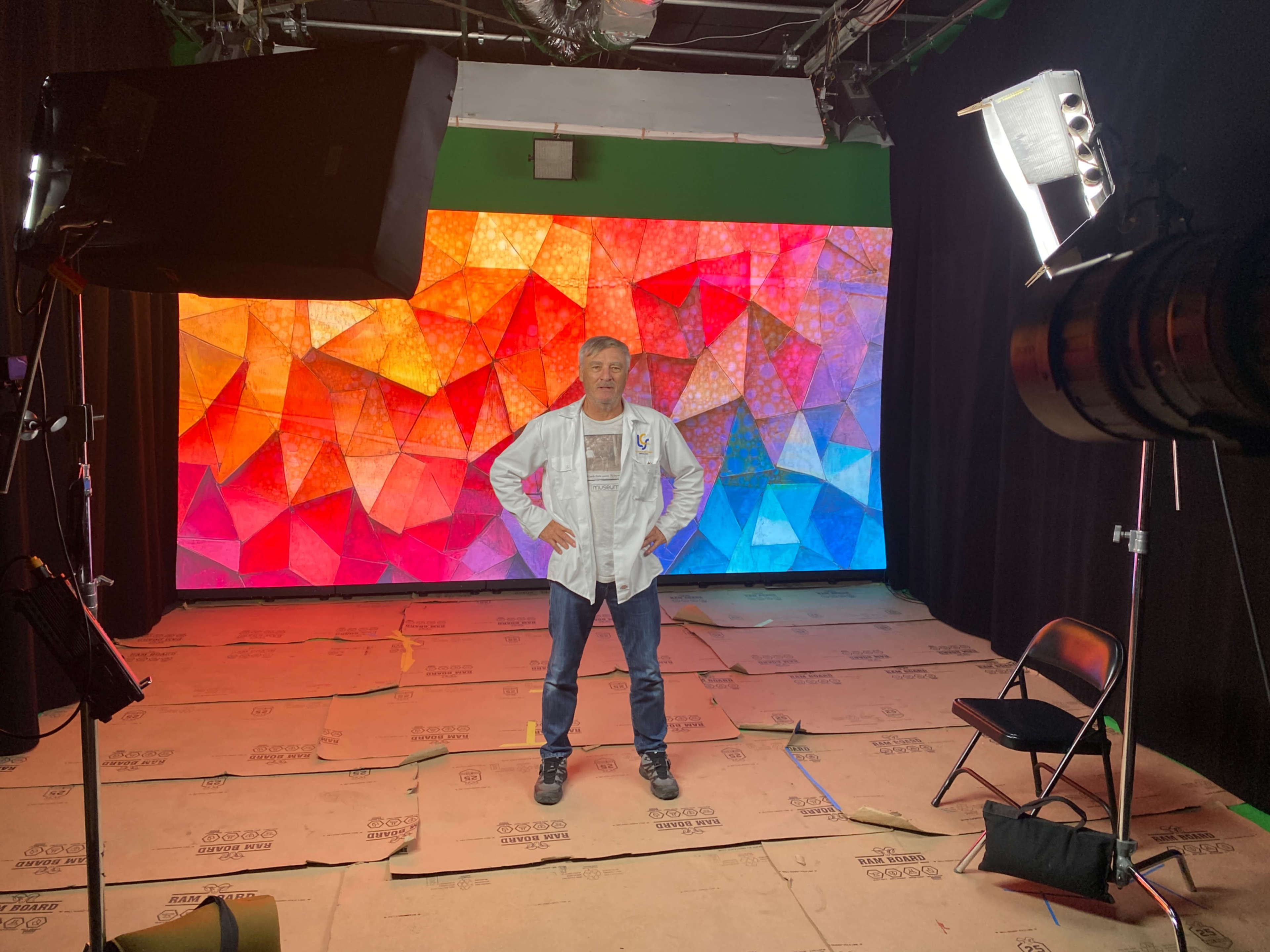 A man in a white jacket stands in front of a colorful geometric backdrop inside a studio setting.