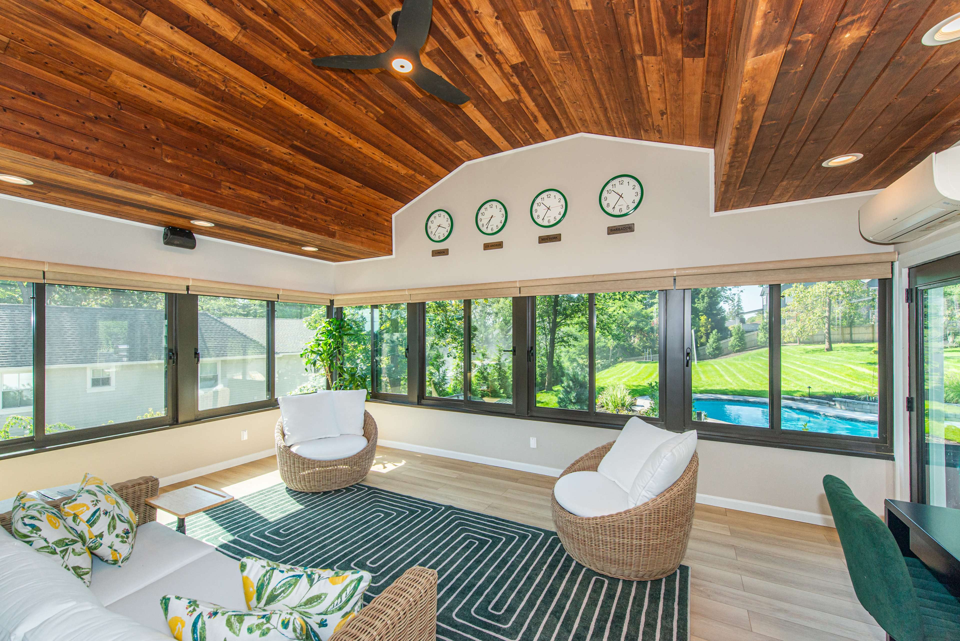 A sunroom features large windows, two chairs with cushions, and several clocks on the wall, with a view of a green landscape outside.