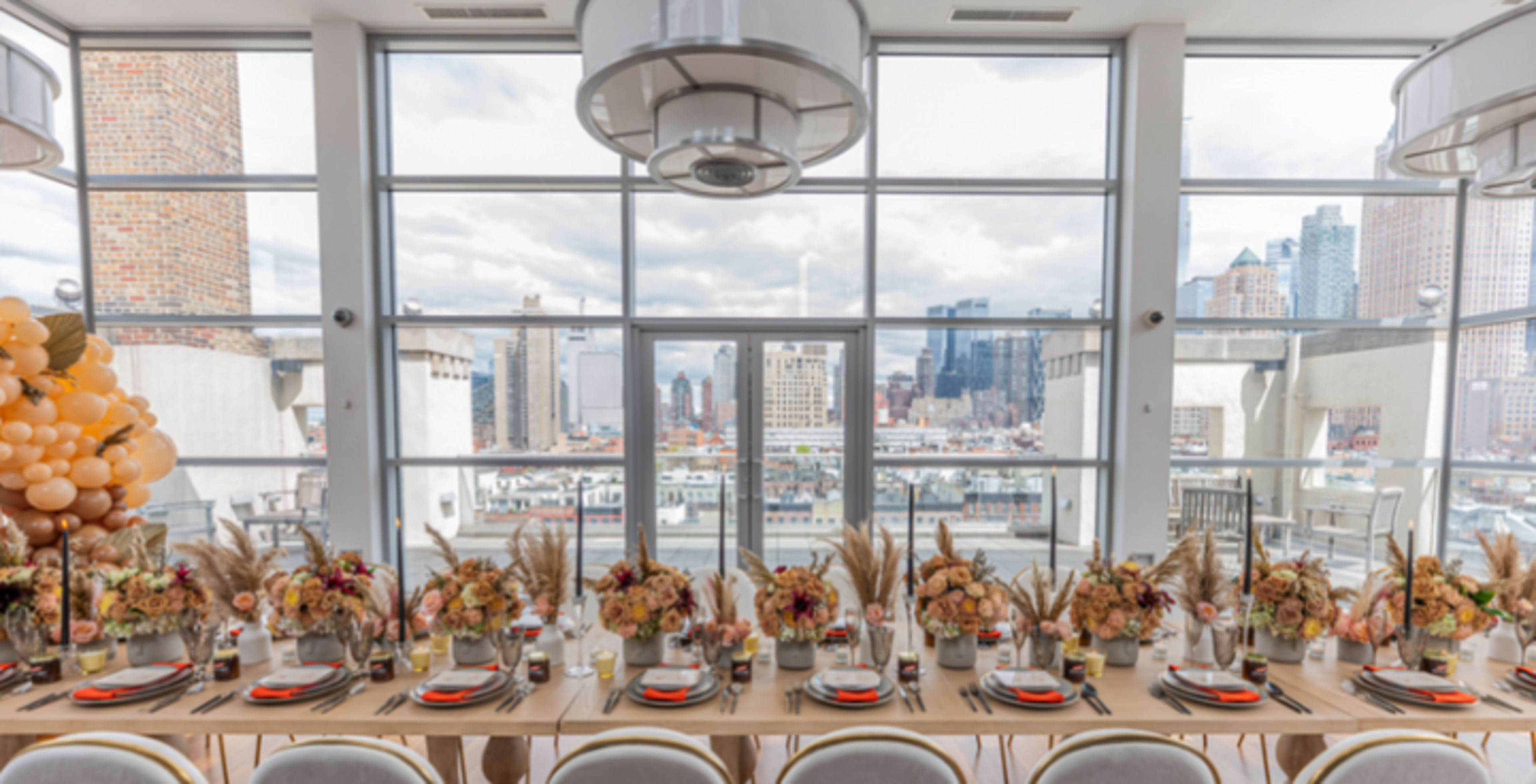 A long dining table is set with floral arrangements and tableware, situated against large windows that showcase a city skyline.