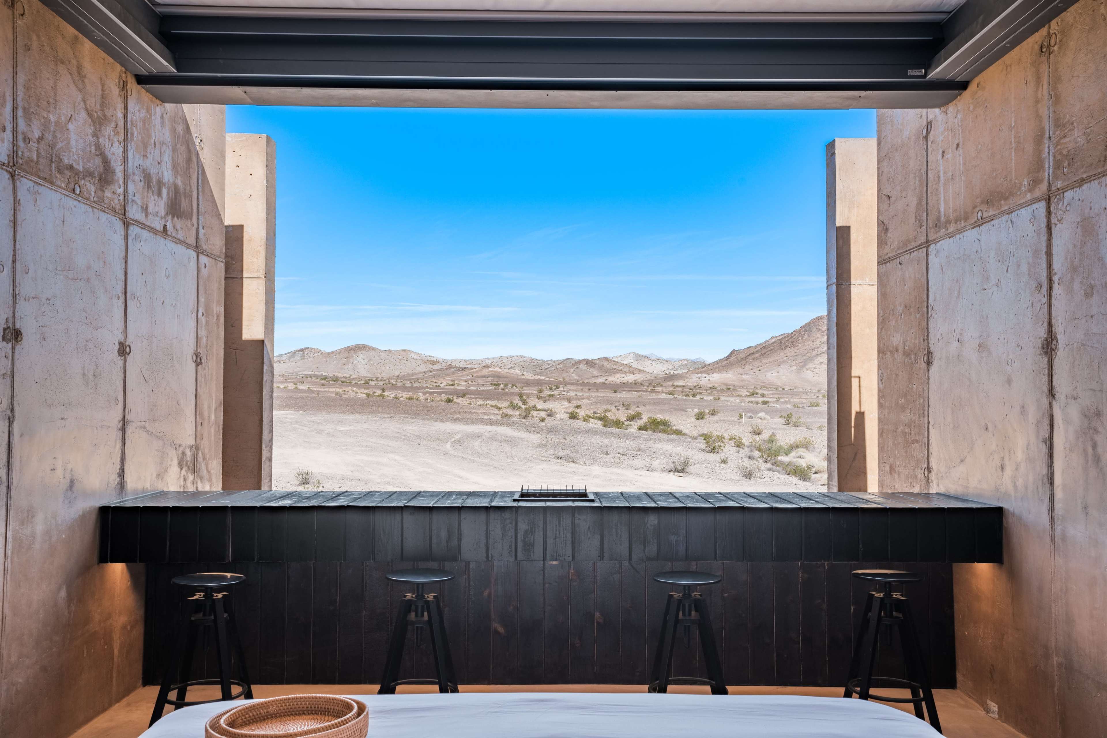 A minimalist interior with a black bar and stools overlooking a expansive desert landscape framed by concrete walls.