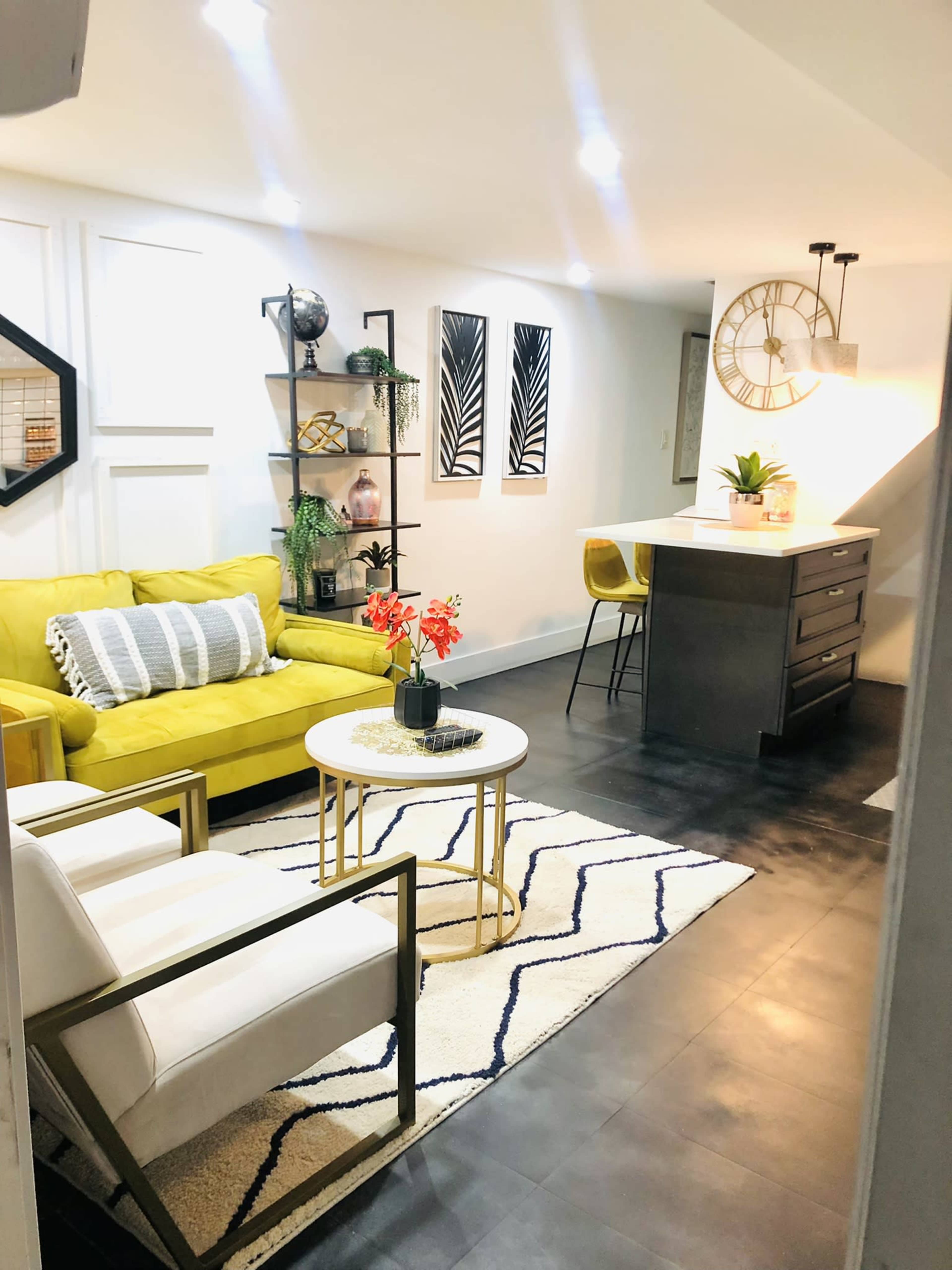 The image shows a modern living room with a yellow sofa, two white chairs, a round coffee table, and a dining area featuring a bar counter and stylish decor.