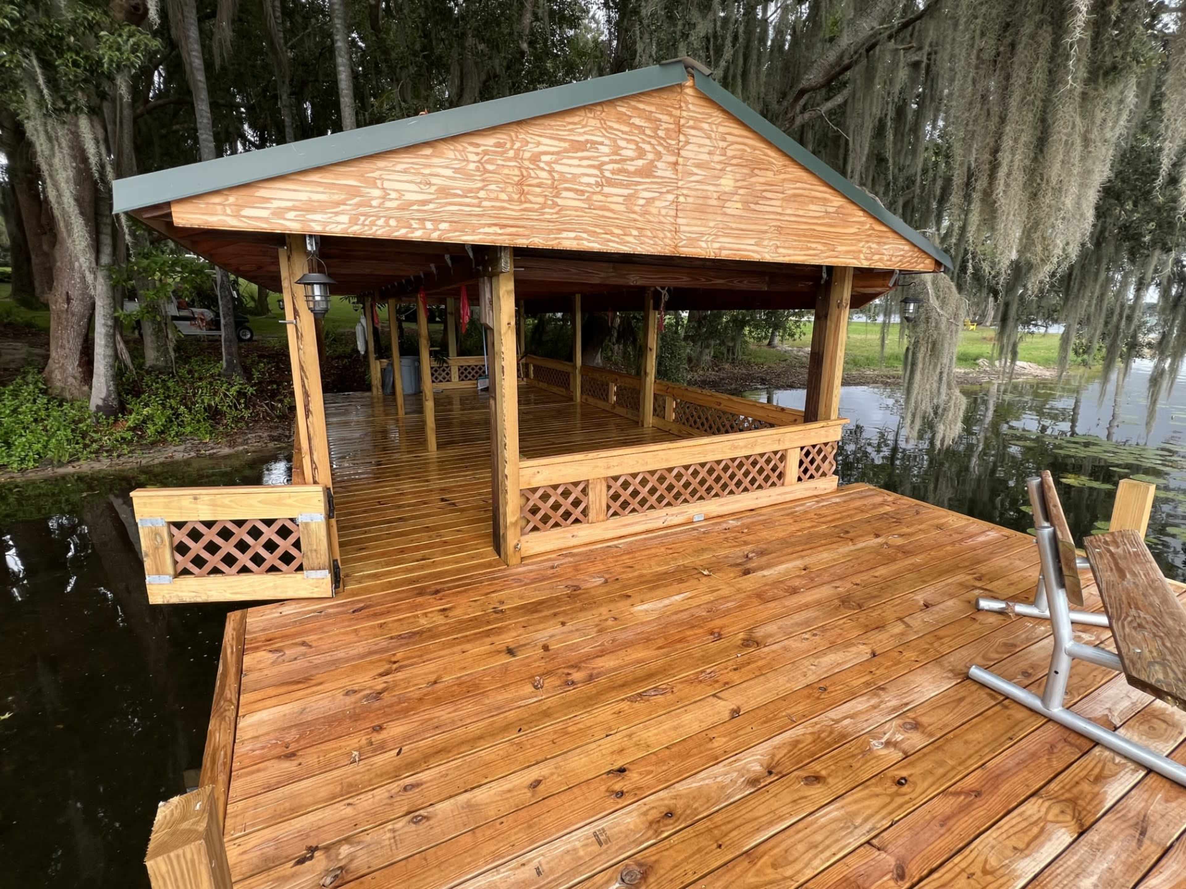 A wooden dock with a covered pavilion extends over a calm body of water, surrounded by trees.