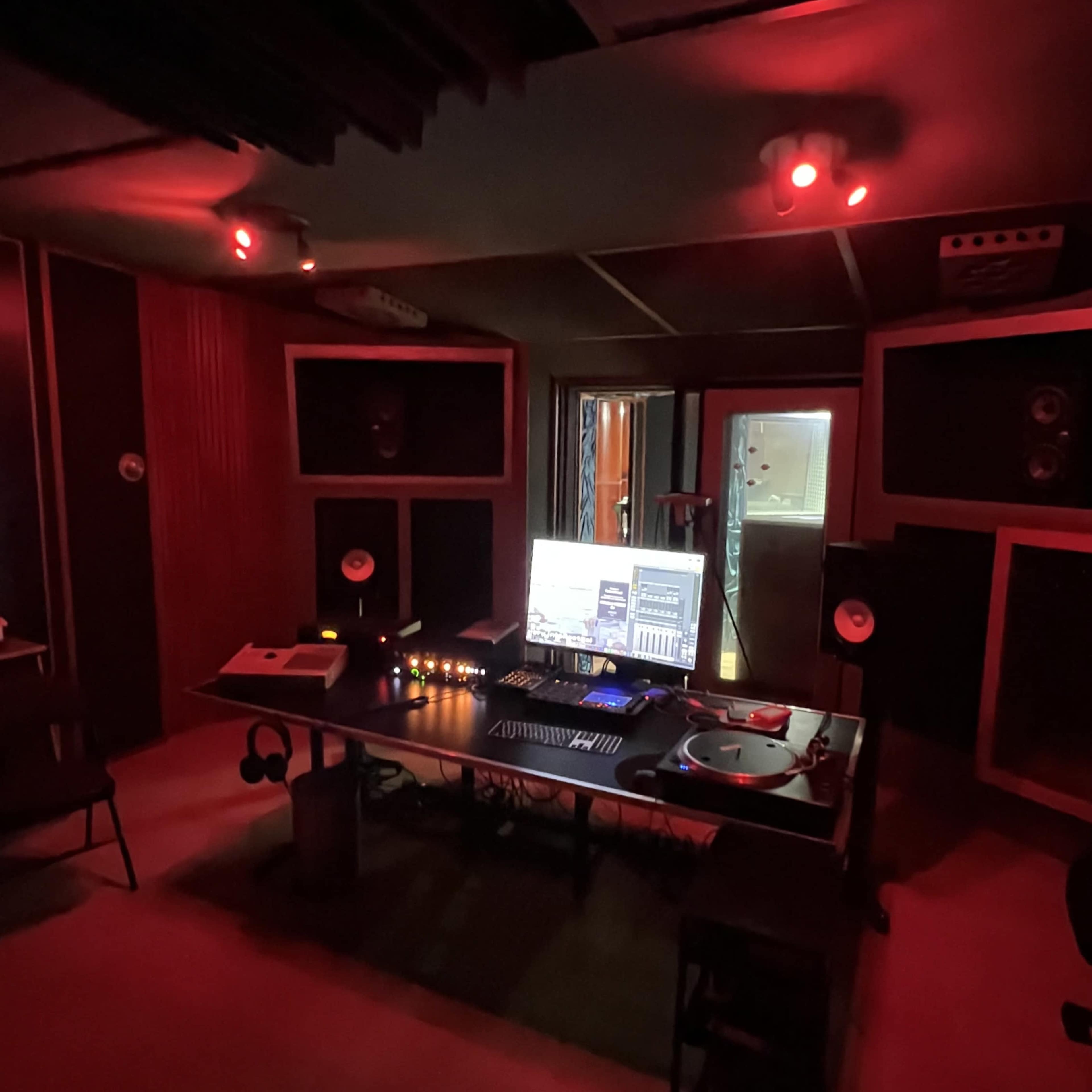 The image shows a recording studio with red lighting, featuring a desk with audio equipment, a computer monitor, and soundproof walls.