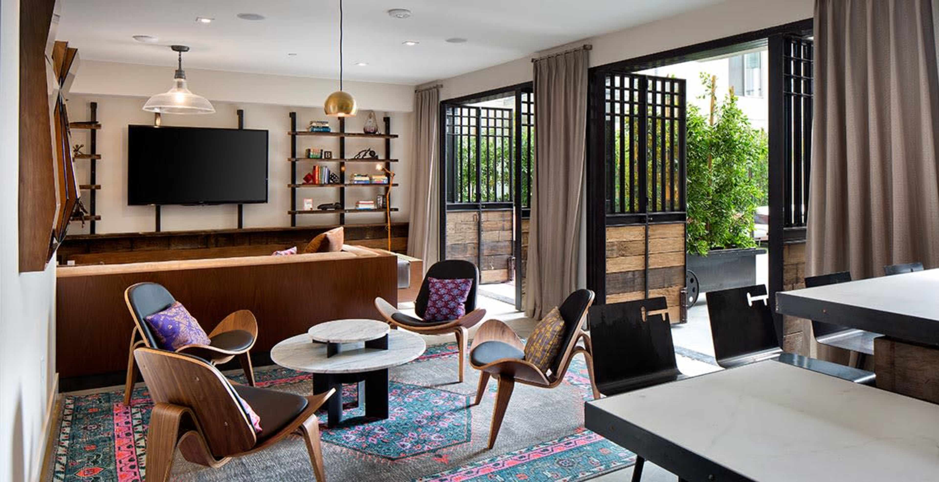 The image shows a modern living room with a television mounted on the wall, comfortable seating arranged around a coffee table, and large windows opening to a patio.