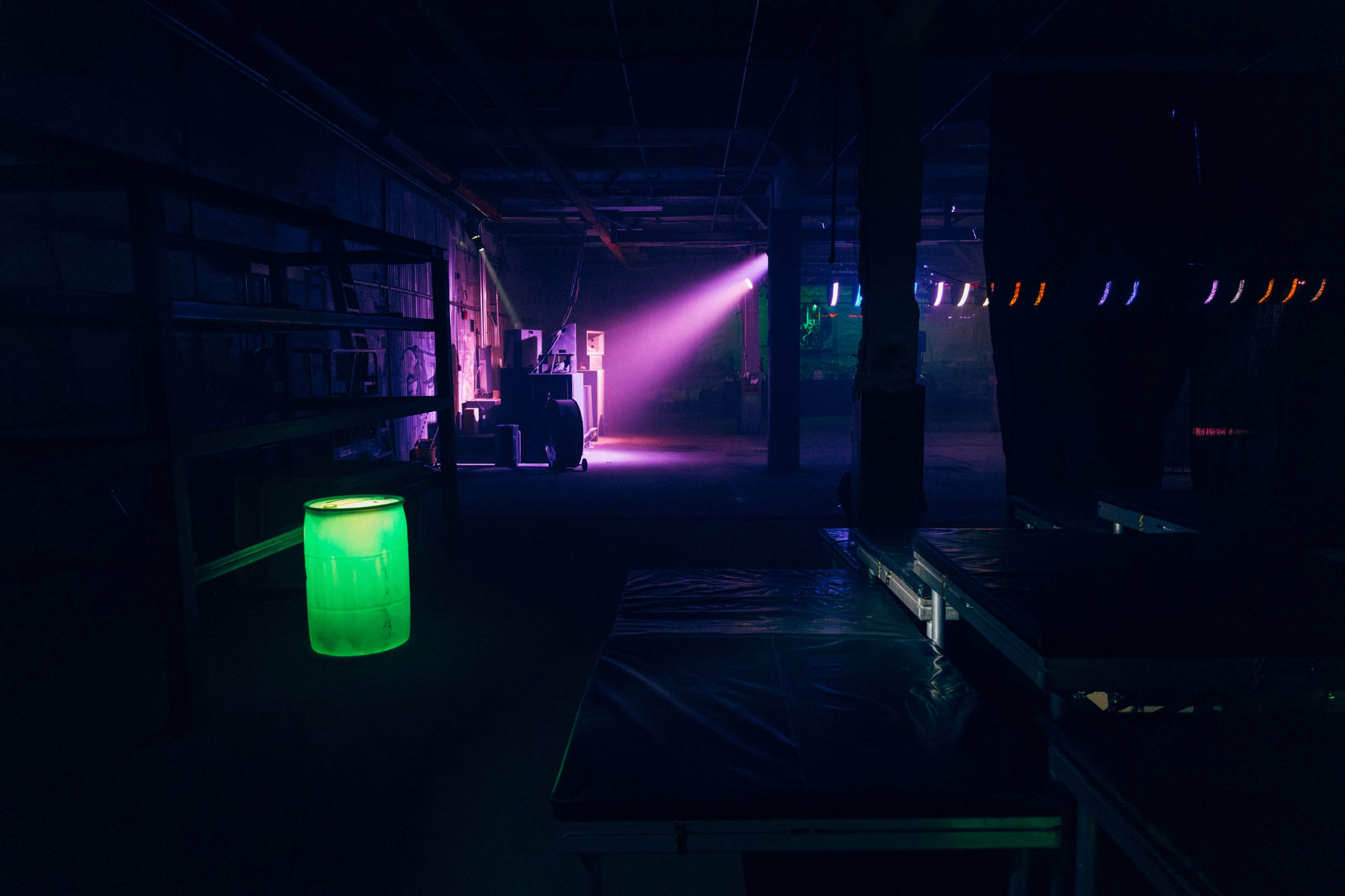 The image shows a dimly lit indoor space with a glowing green barrel and colorful light beams illuminating the area.